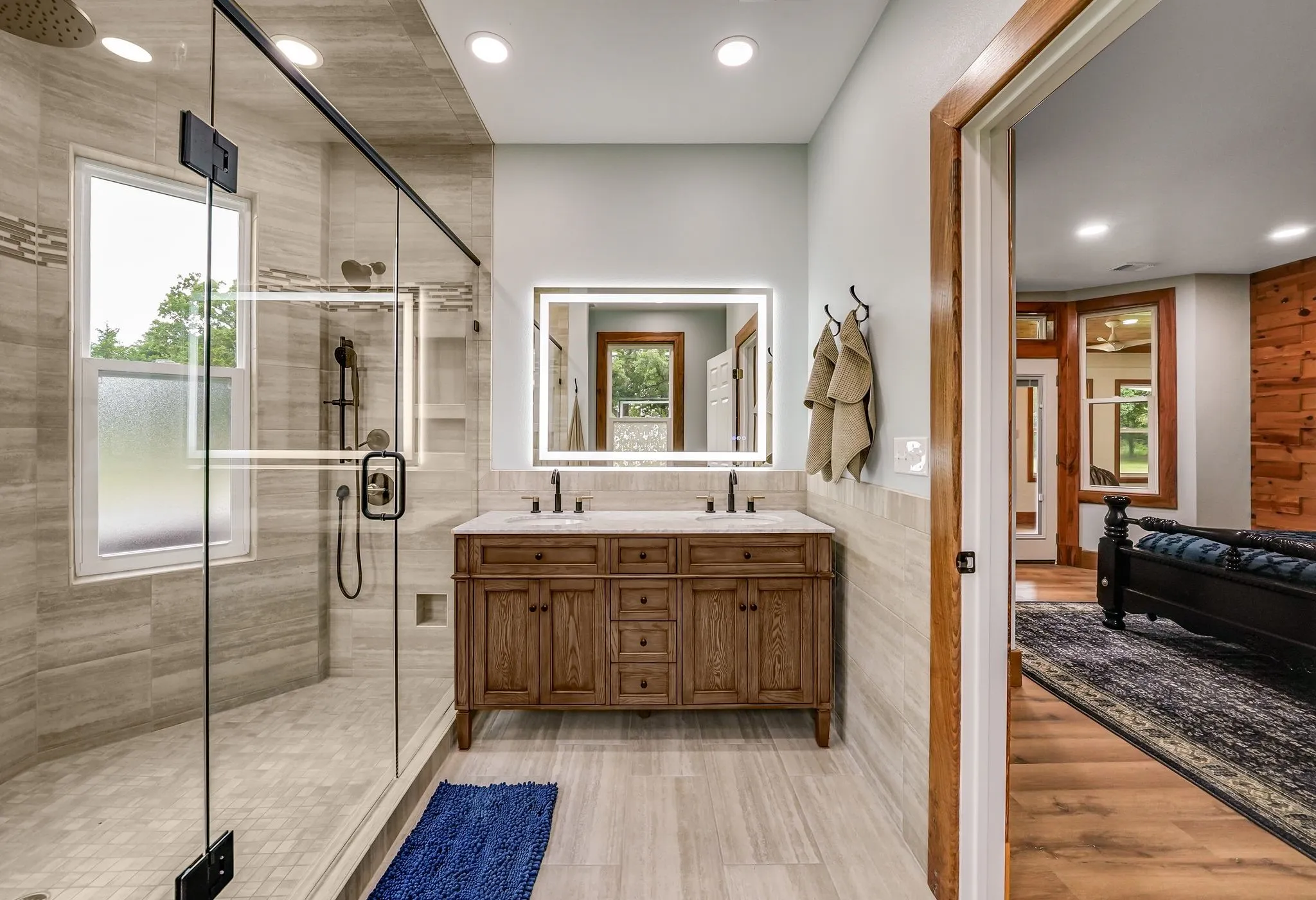 Spacious bathroom featuring a frameless glass shower enclosure, a dual vanity with a light-colored countertop, an illuminated mirror, and natural wood-finish cabinetry