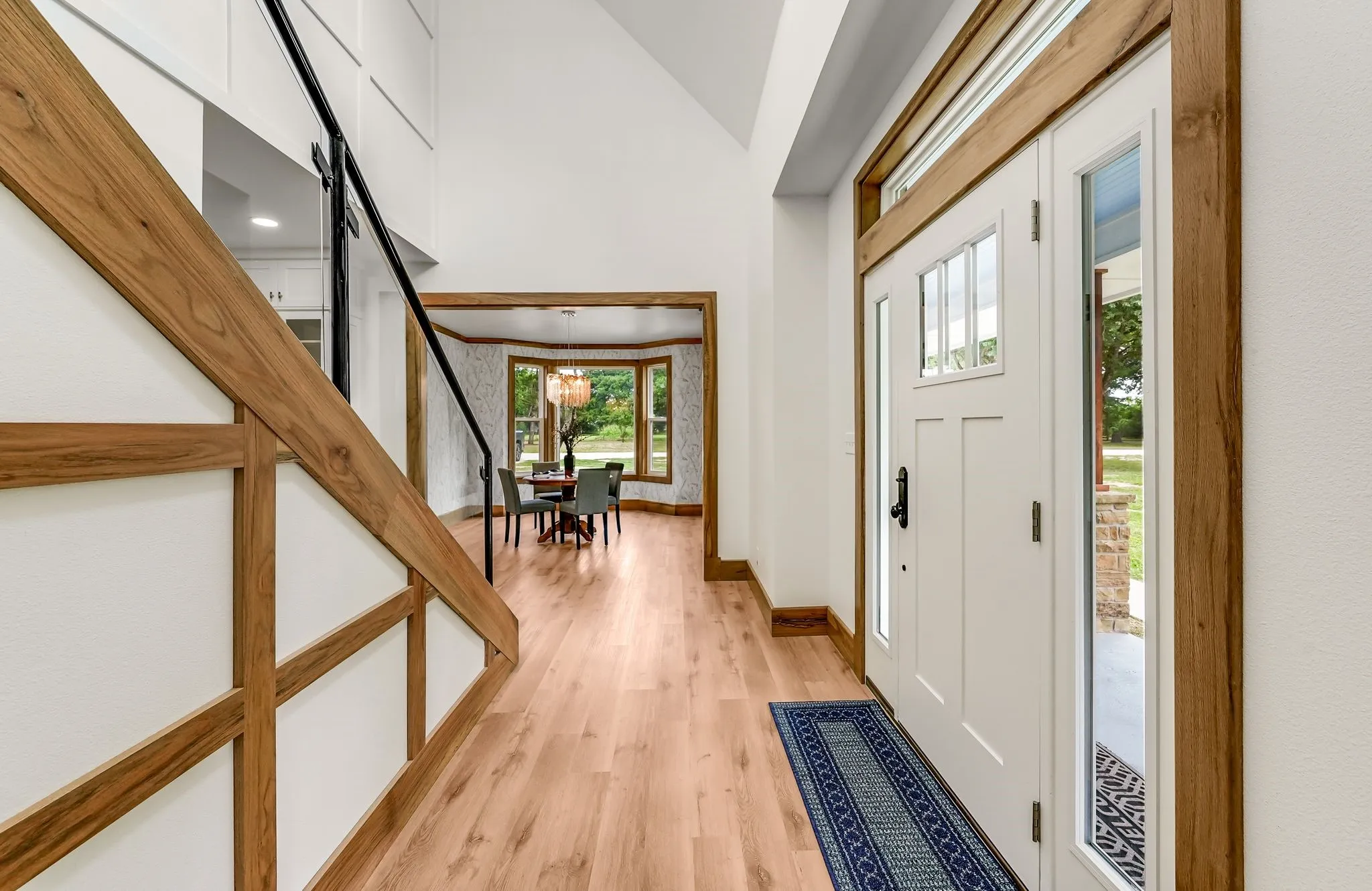 Entry foyer featuring a white craftsman-style door with sidelights and transom window, light wood-finish flooring, a wood and black metal staircase, vaulted ceilings, and a pass-through to an additional room