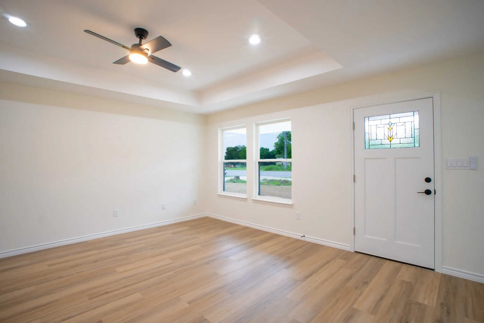 Open living area featuring wood-finish flooring, a tray ceiling with recessed lighting and a ceiling fan, and a white entry door with stained glass insert