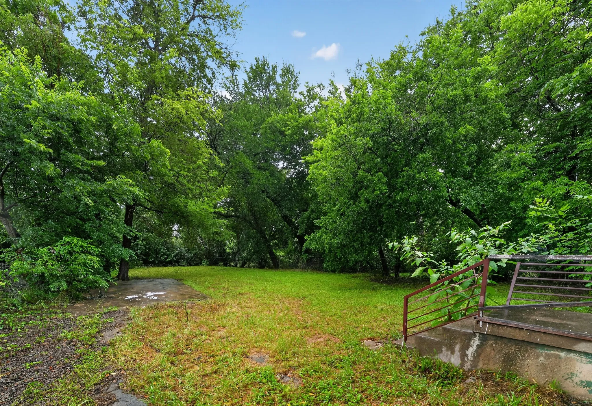 Expansive grassy area surrounded by mature trees, featuring a concrete pad and a concrete stair structure with metal railings
