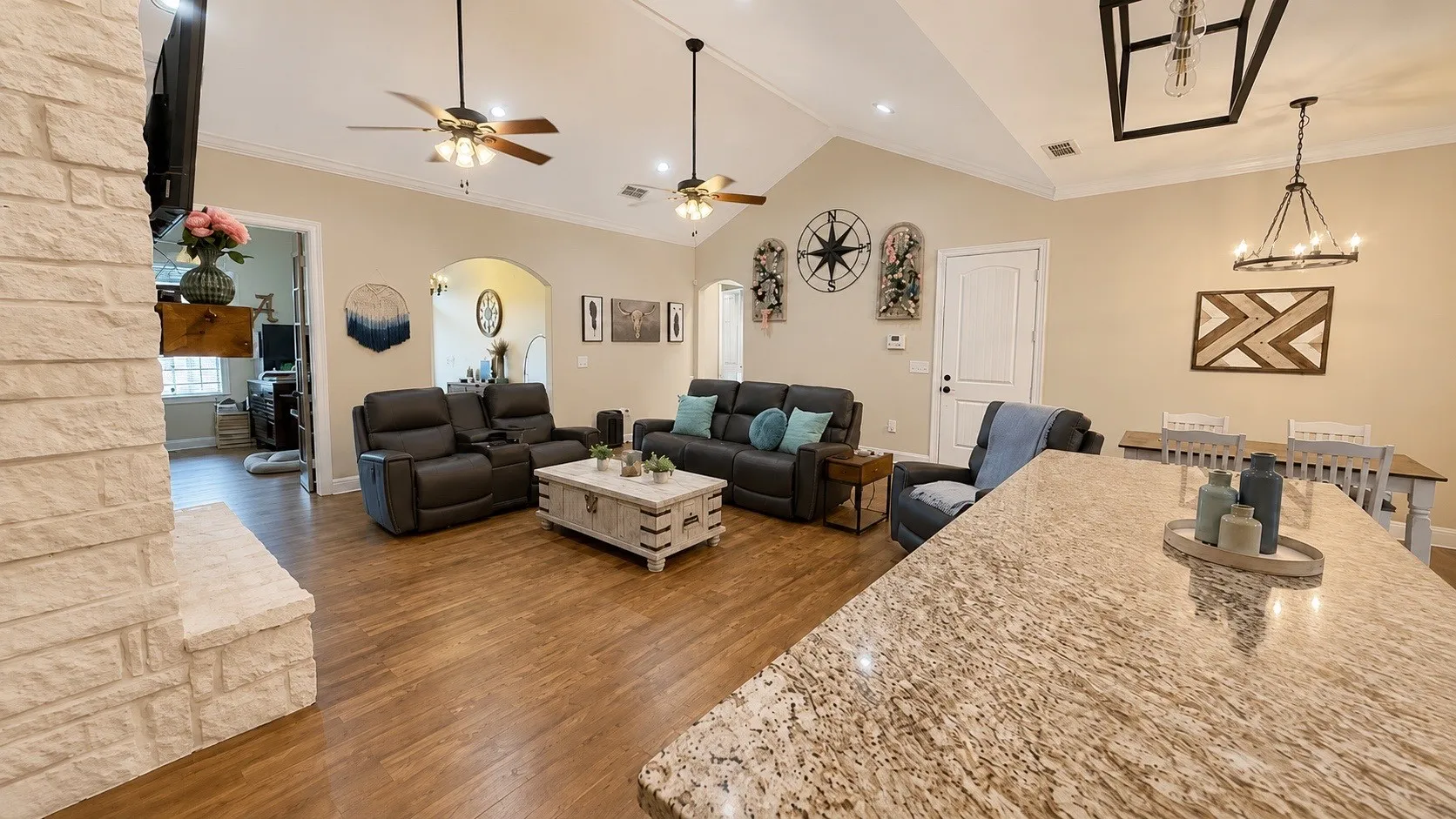 Spacious living area featuring vaulted ceilings with exposed beams, wood-finish flooring, a stone-clad fireplace, and a large kitchen island with a granite countertop