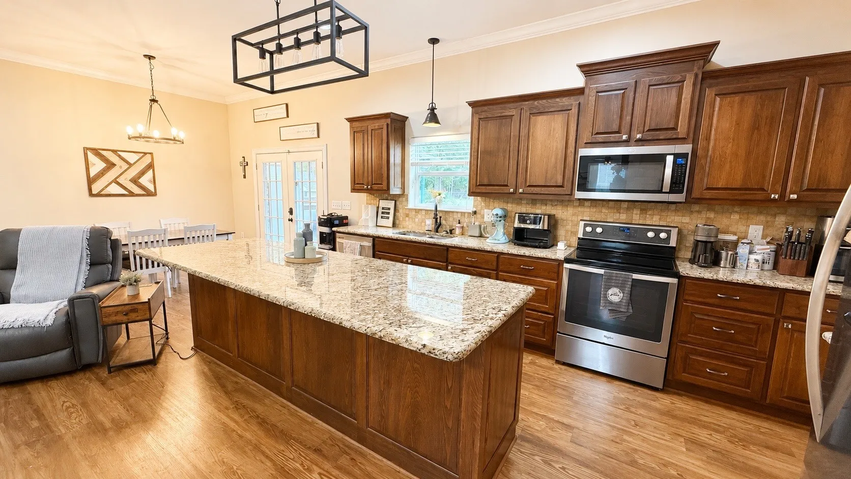 Open-concept kitchen featuring an oversized island with a light-toned countertop, wood-finish flooring, full-height wood cabinetry, stainless steel appliances, and a tiled backsplash
