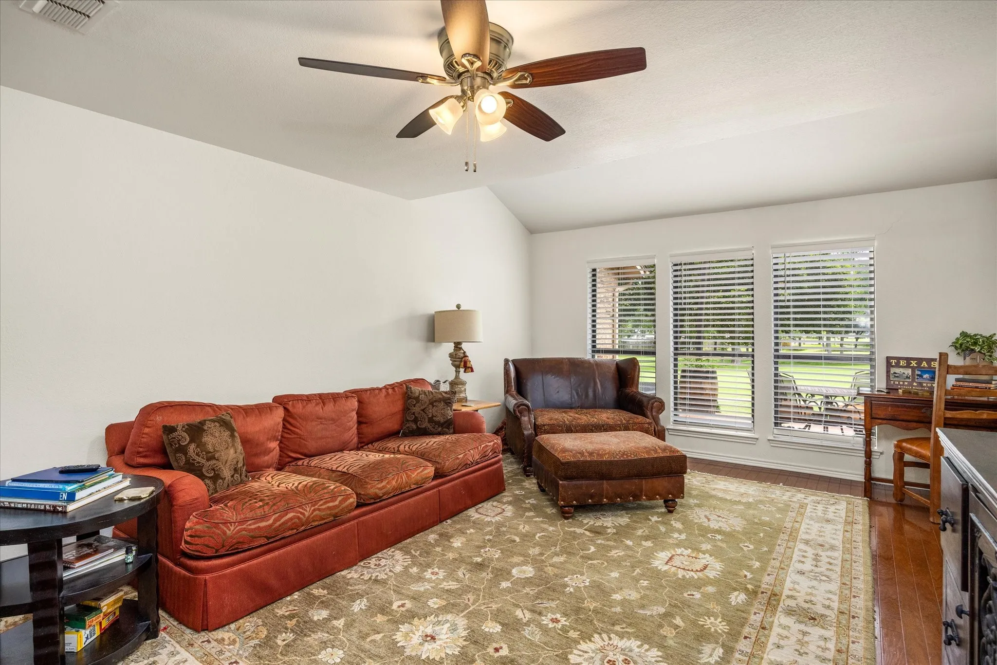 Living area featuring vaulted ceilings, wood-finish flooring, a ceiling fan with integrated lighting, and a trio of windows providing natural illumination