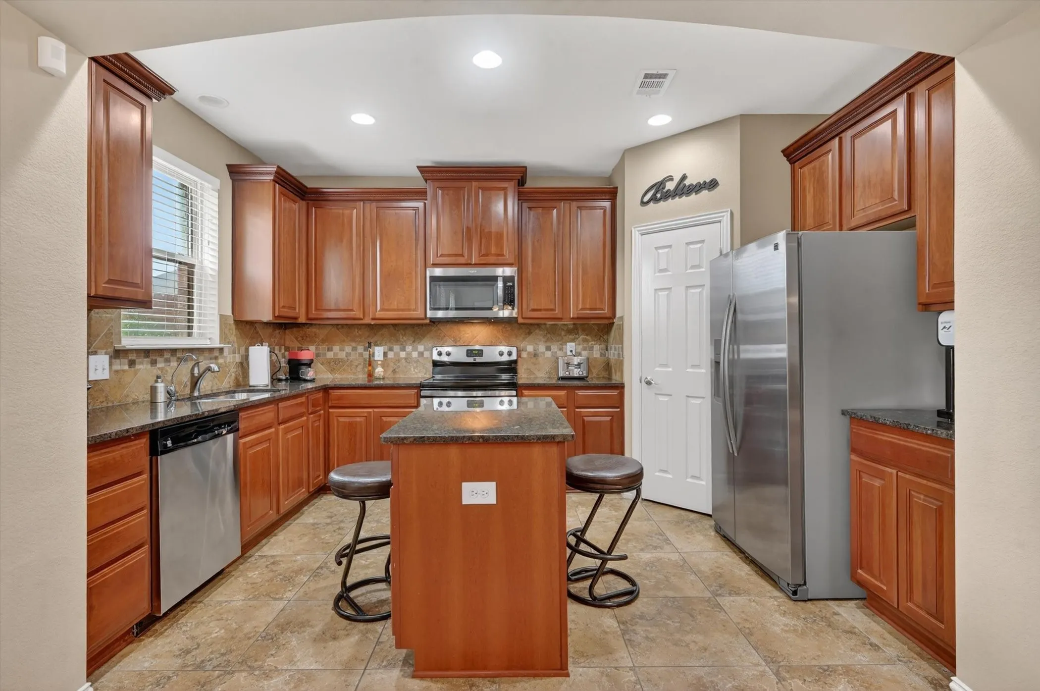 Spacious kitchen featuring a central island with seating, dark stone countertops, wood cabinetry, stainless steel appliances, and tile flooring