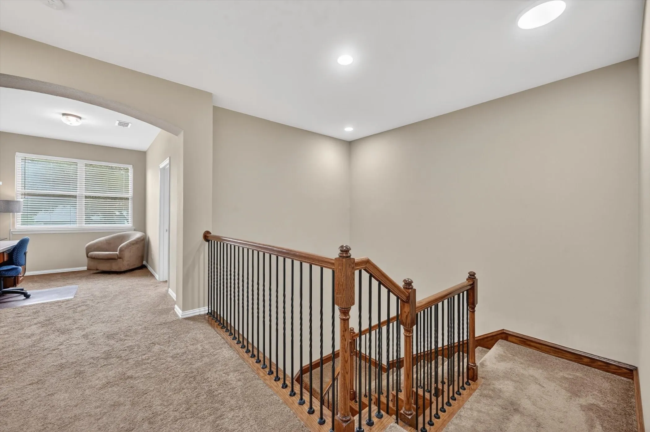 Carpeted landing featuring a wood and wrought iron staircase railing, recessed lighting, and an arched entry to an additional carpeted room with multiple windows