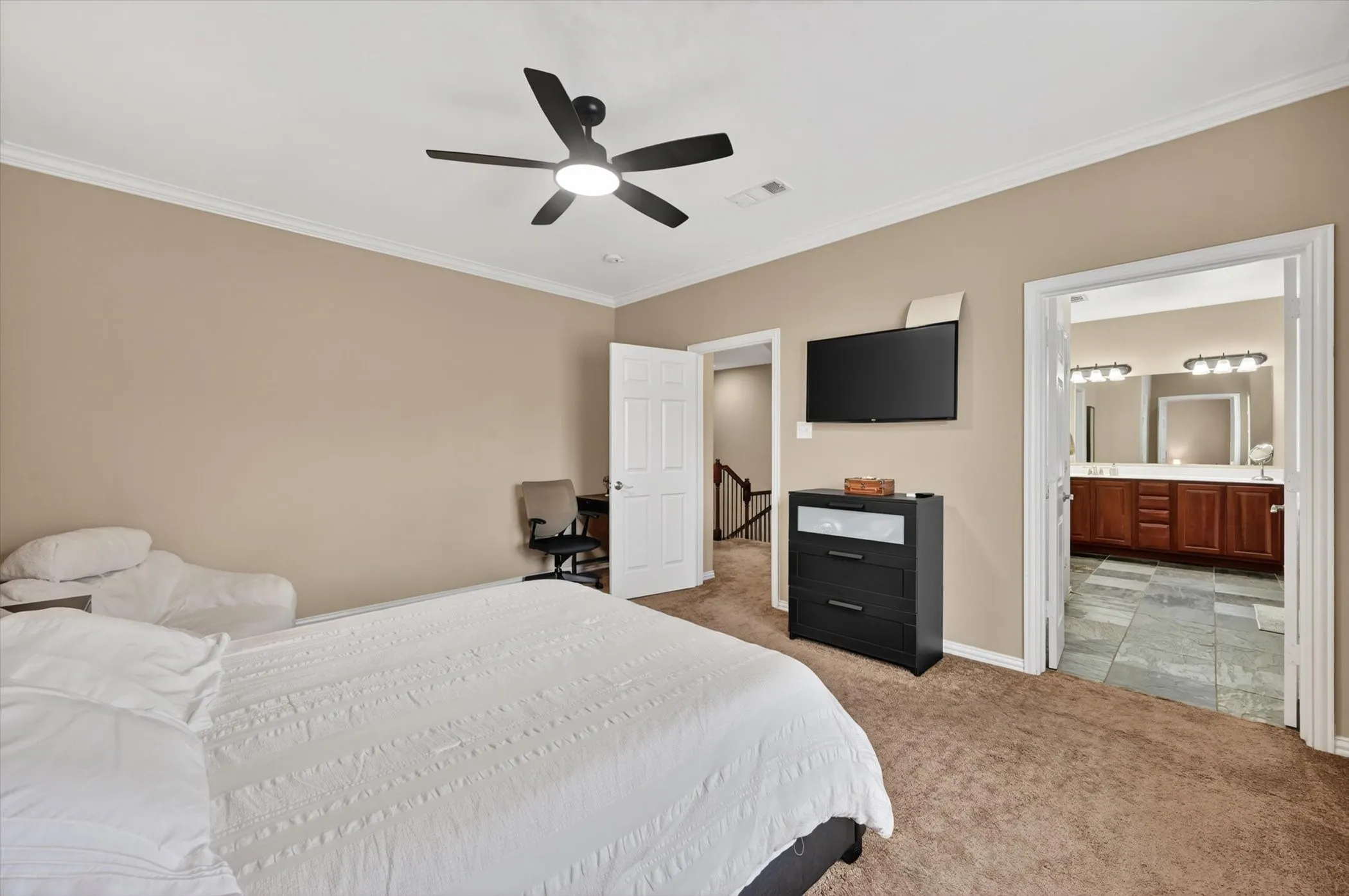 Carpeted bedroom featuring a ceiling fan with integrated lighting, crown molding, and direct entry to a bathroom with dual vanity sinks and a tiled floor