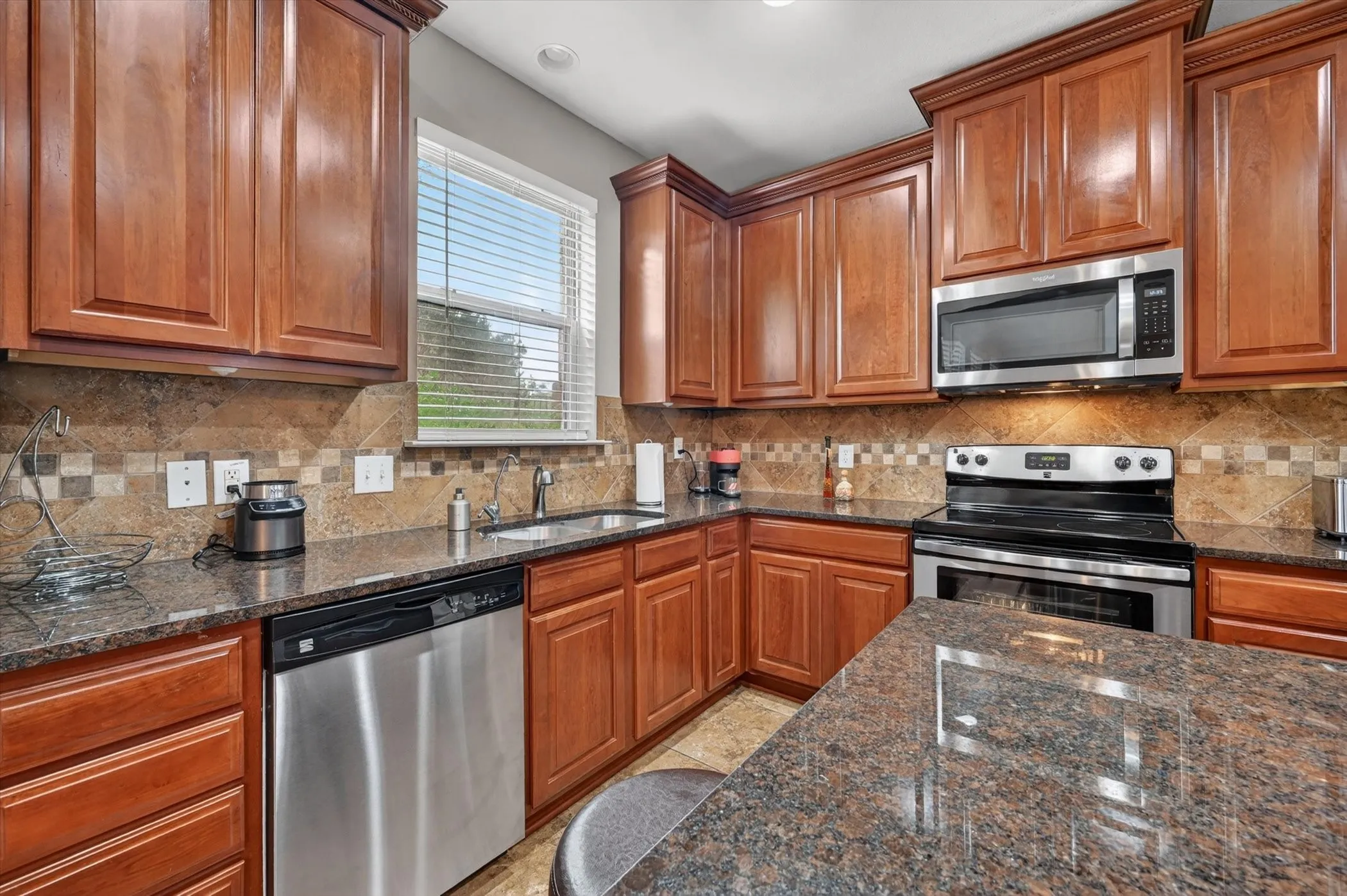Kitchen featuring wood cabinetry, stone countertops, stainless steel appliances, a tile backsplash, and a window with blinds