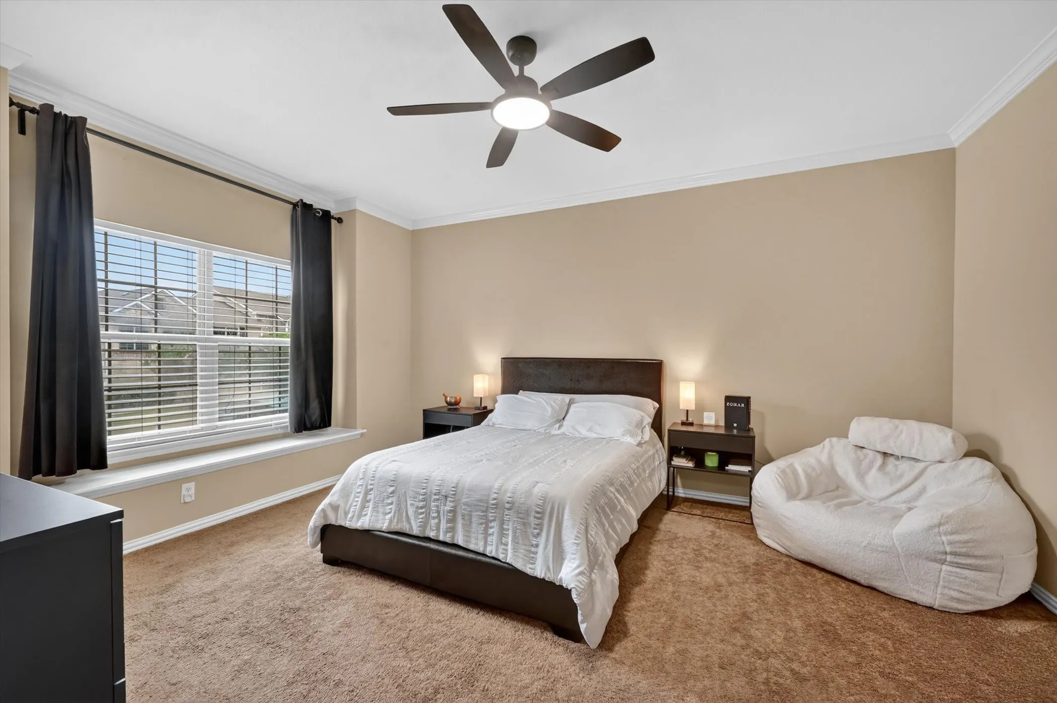 Spacious bedroom featuring neutral toned walls, brown carpet flooring, a ceiling fan with integrated lighting, and a large window with white blinds and dark curtain rods