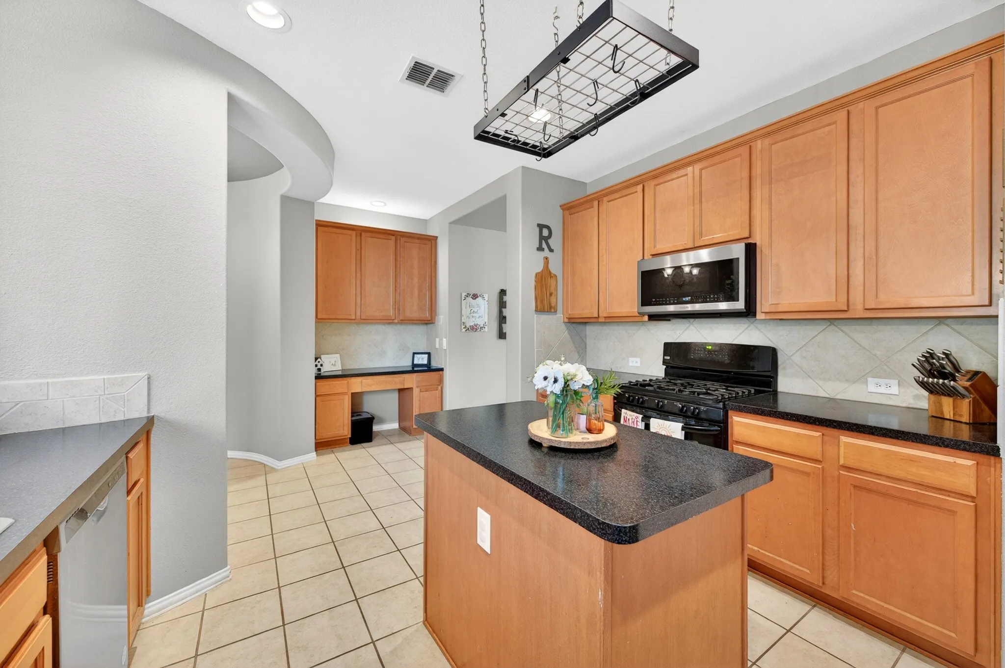 Kitchen with a central island featuring a dark countertop, light wood-finish cabinetry, stainless steel microwave, black gas range, and tile flooring