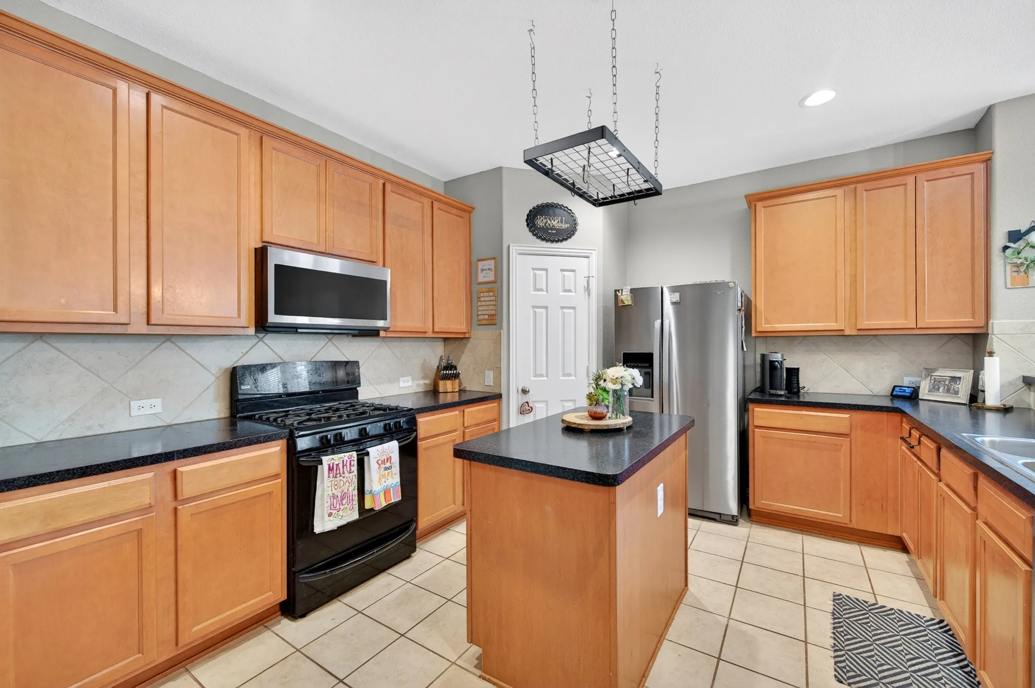 Kitchen featuring an island, light wood cabinetry, dark countertops, stainless steel refrigerator, and a black range with an over-the-range microwave