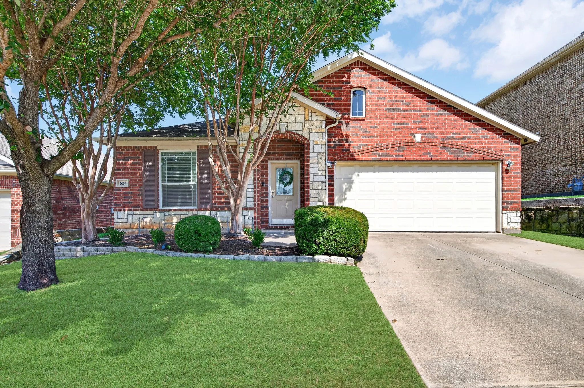 Brick and stone facade featuring a two-car garage, mature trees, and landscaping
