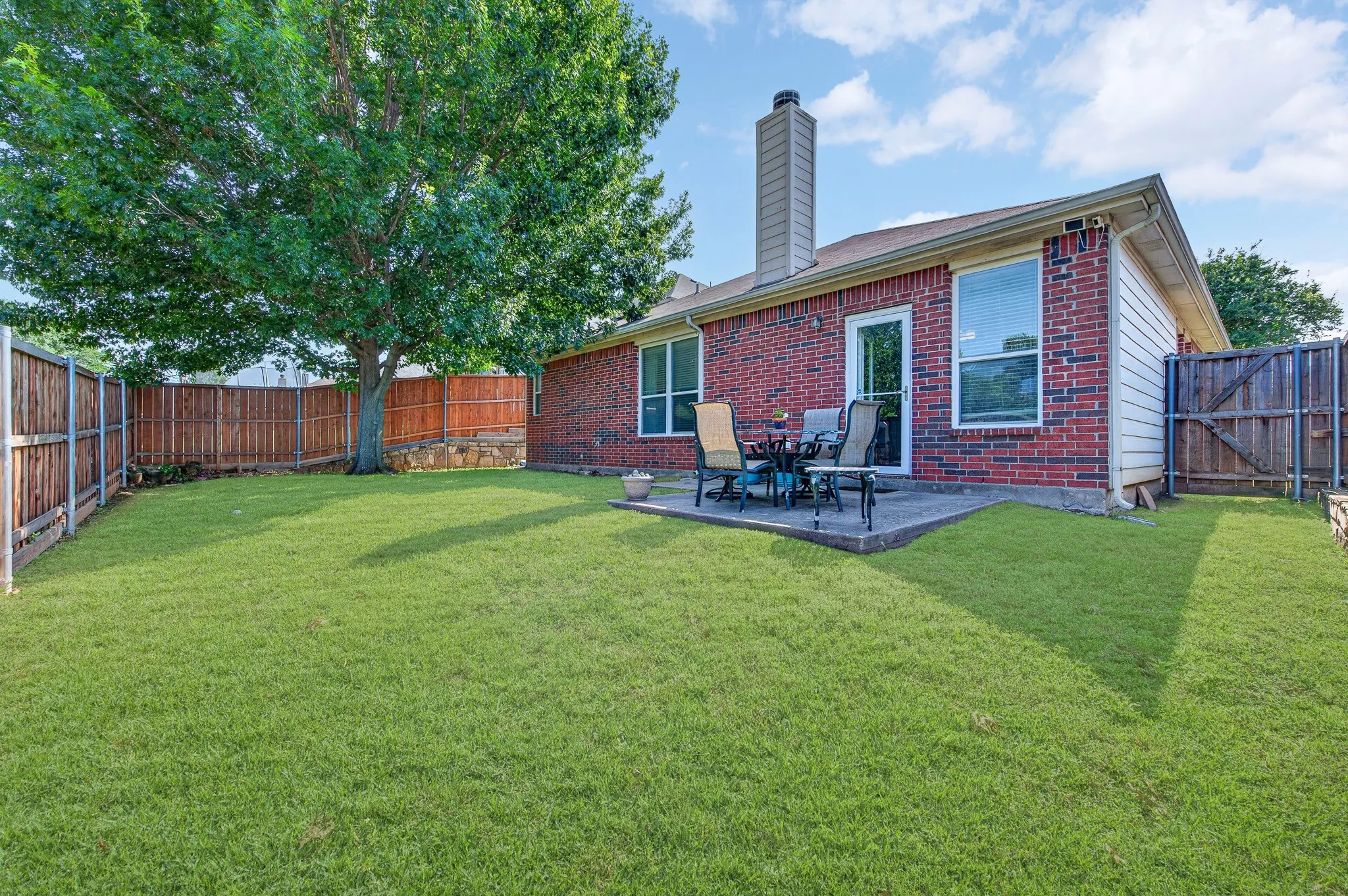 Expansive backyard featuring a manicured lawn, a mature shade tree, a brick exterior, a concrete patio, and a wood privacy fence