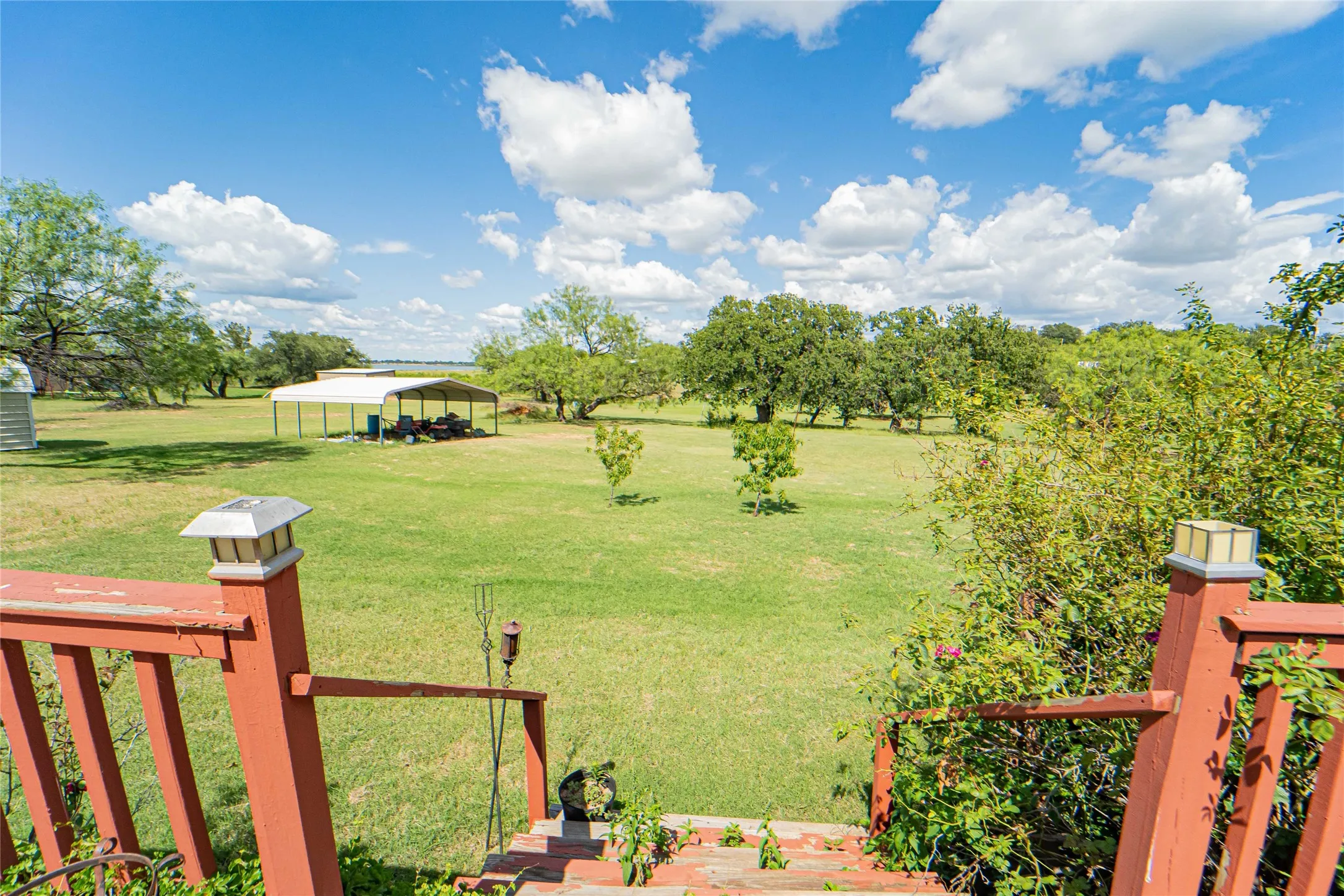 Expansive green lawn extending to a distant body of water, featuring mature trees and a metal carport structure