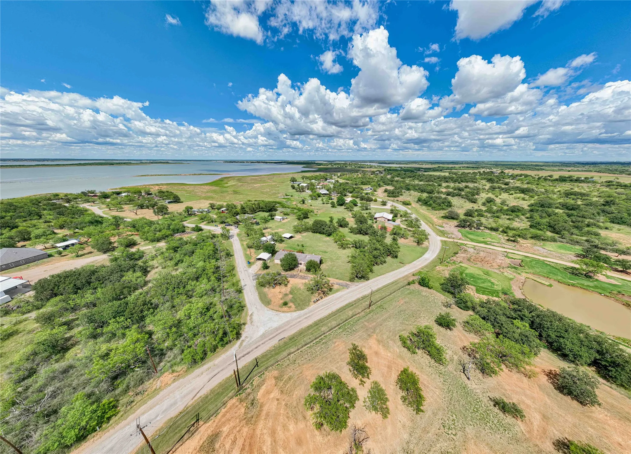 Expansive lakefront property featuring a broad body of water, extensive natural landscaping, multiple mature trees, and a visible roadway