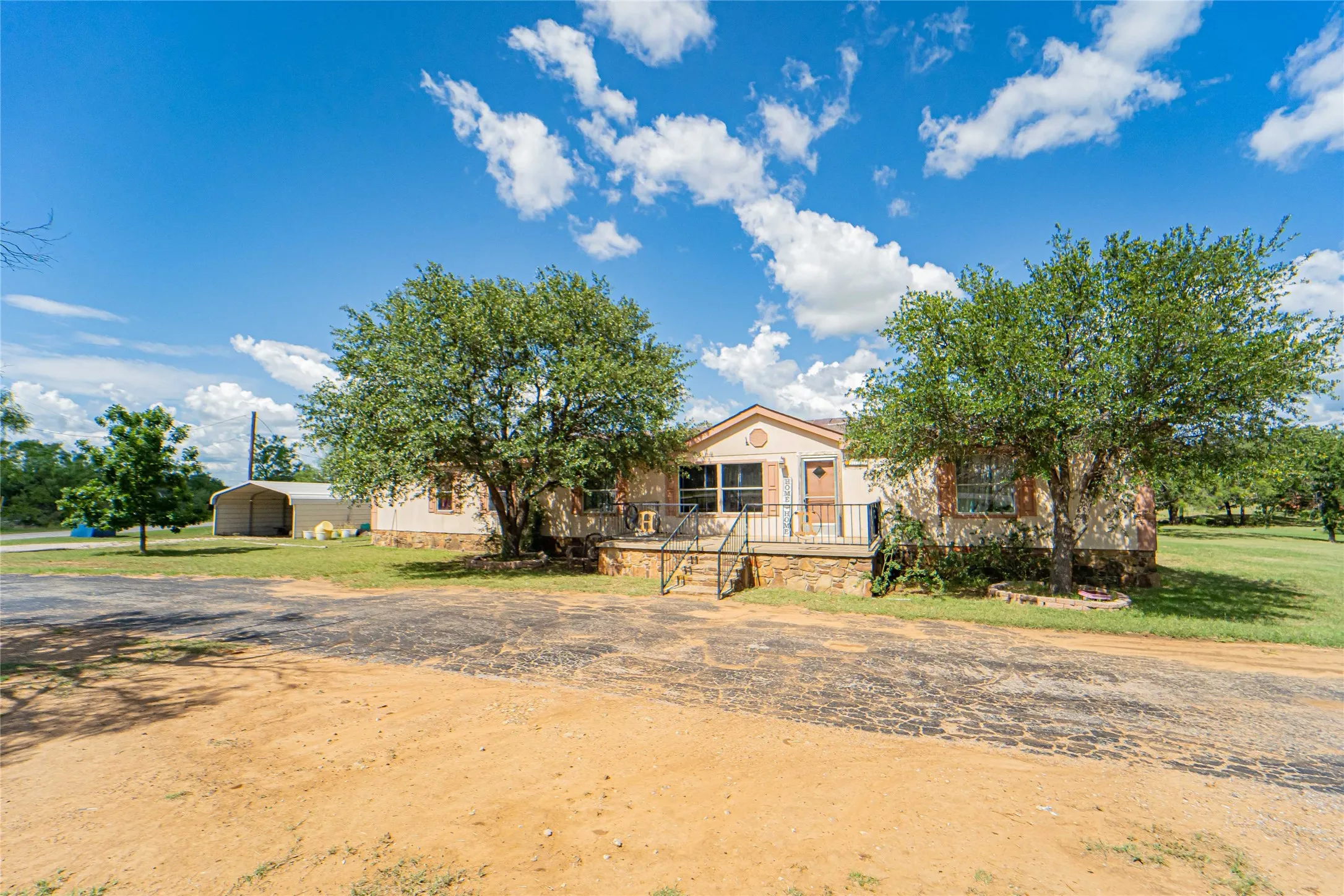 Ranch-style, manufactured home, featuring a covered entry porch with railing, exterior stone detailing, and mature trees