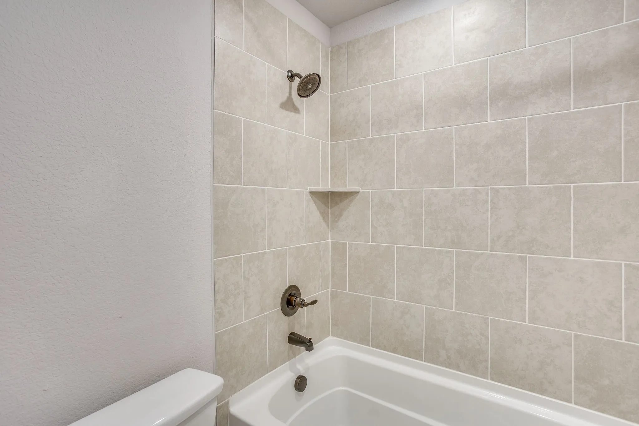 Shower-over-tub combination featuring light-toned rectangular tile surround, oil-rubbed bronze fixtures, and an integrated corner shelf