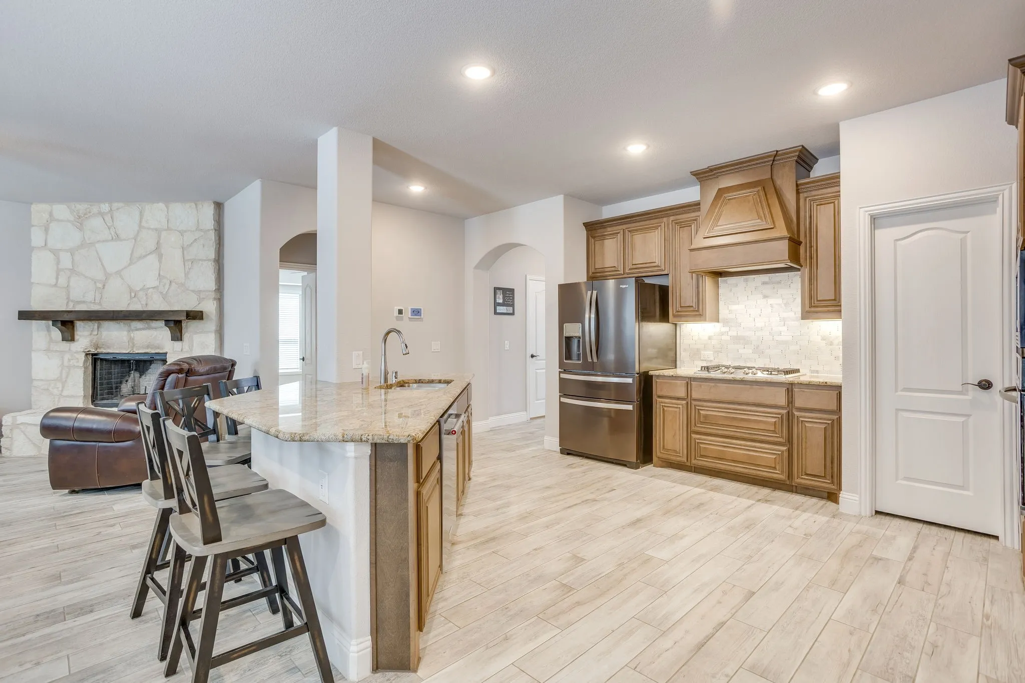 Open-concept living space featuring a central kitchen island with seating, stone fireplace, light wood-finish flooring, recessed lighting, and arched doorways