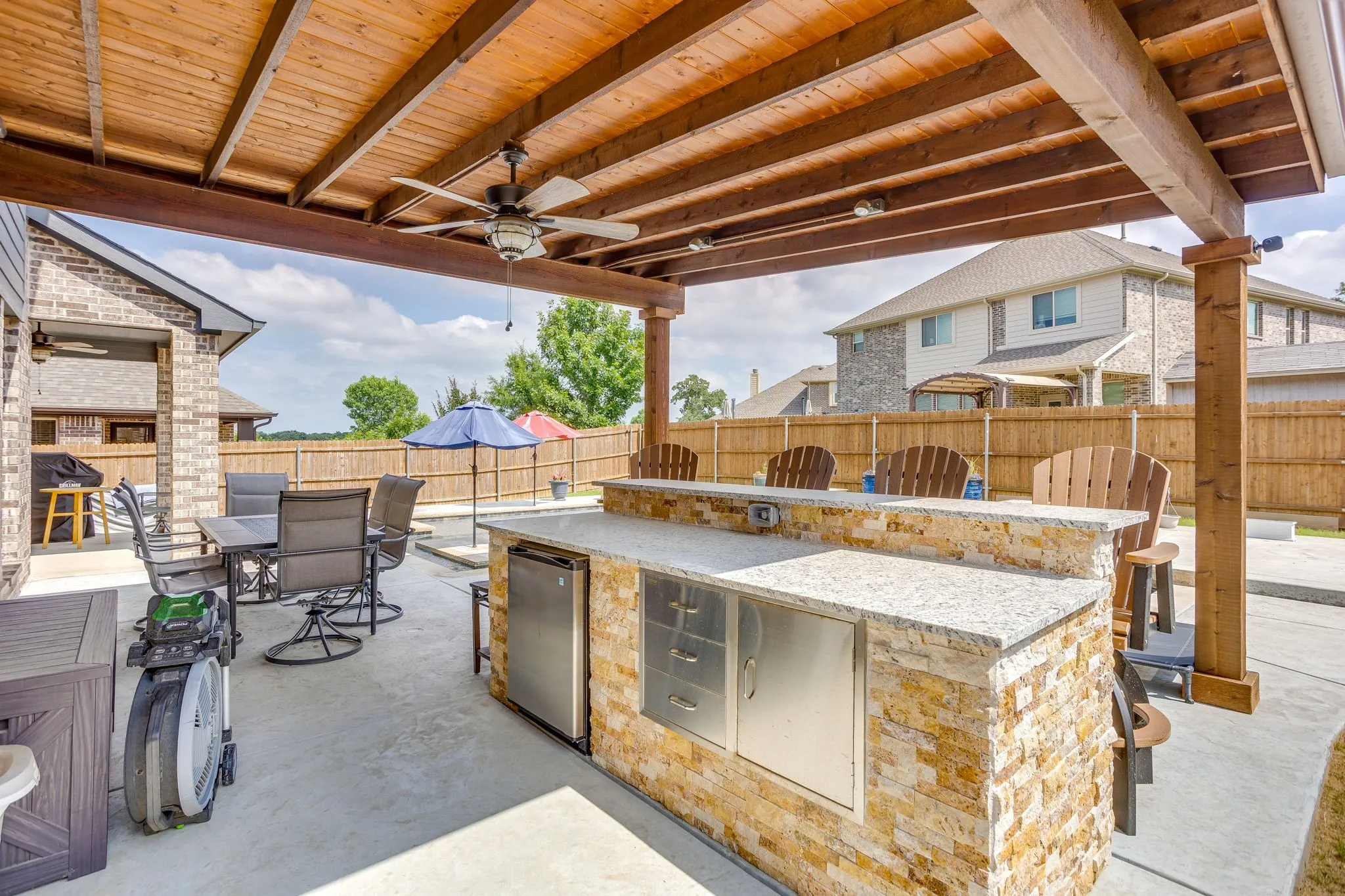 Expansive outdoor living area featuring a stone-clad outdoor kitchen with integrated stainless steel appliances, a wood-finish pergola with a ceiling fan, a paved patio, and a privacy fence