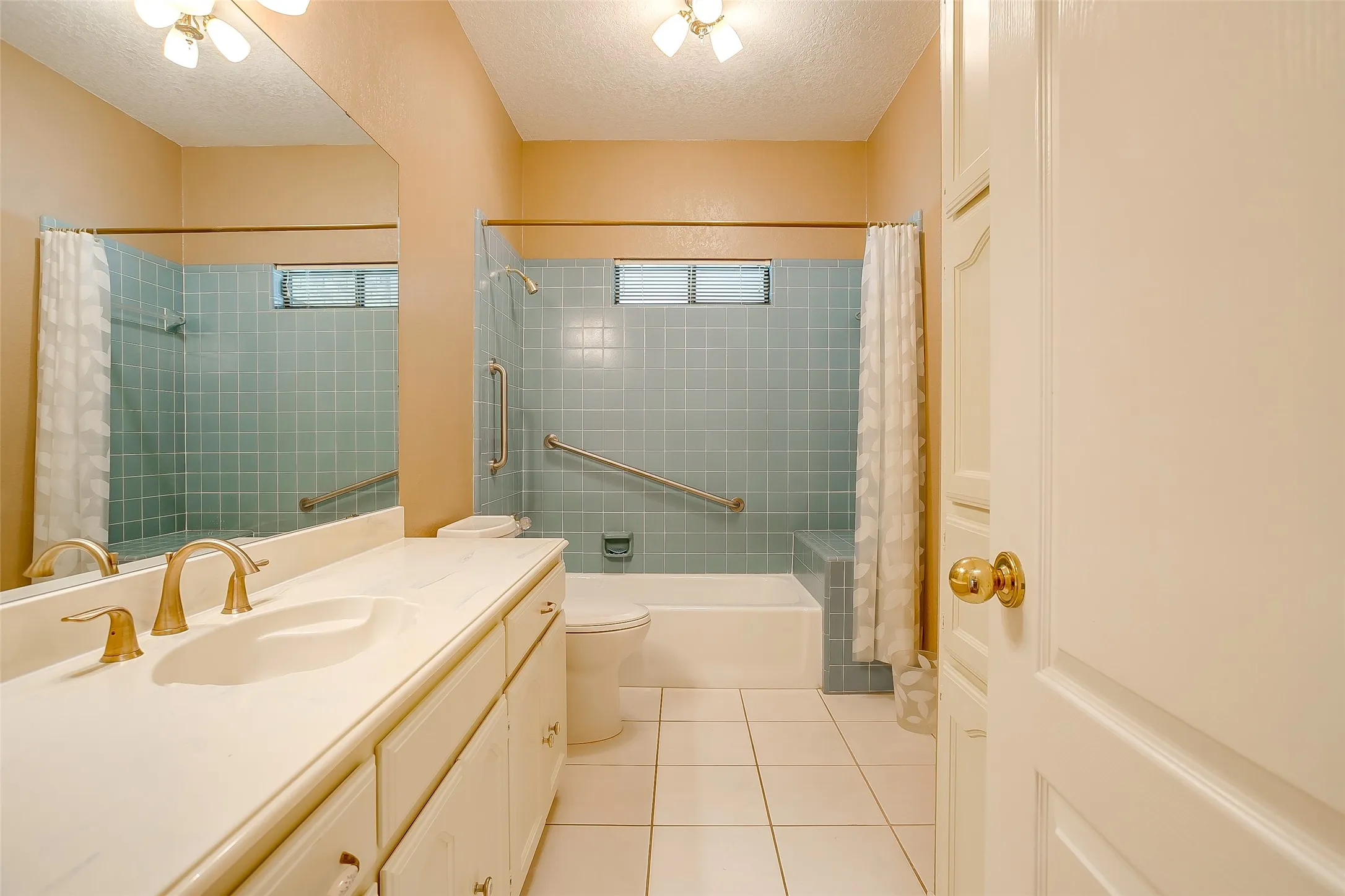 Bathroom featuring a vanity with an integrated sink, gold-tone fixtures, a built-in bathtub with light blue tiled surround, and white floor tiling