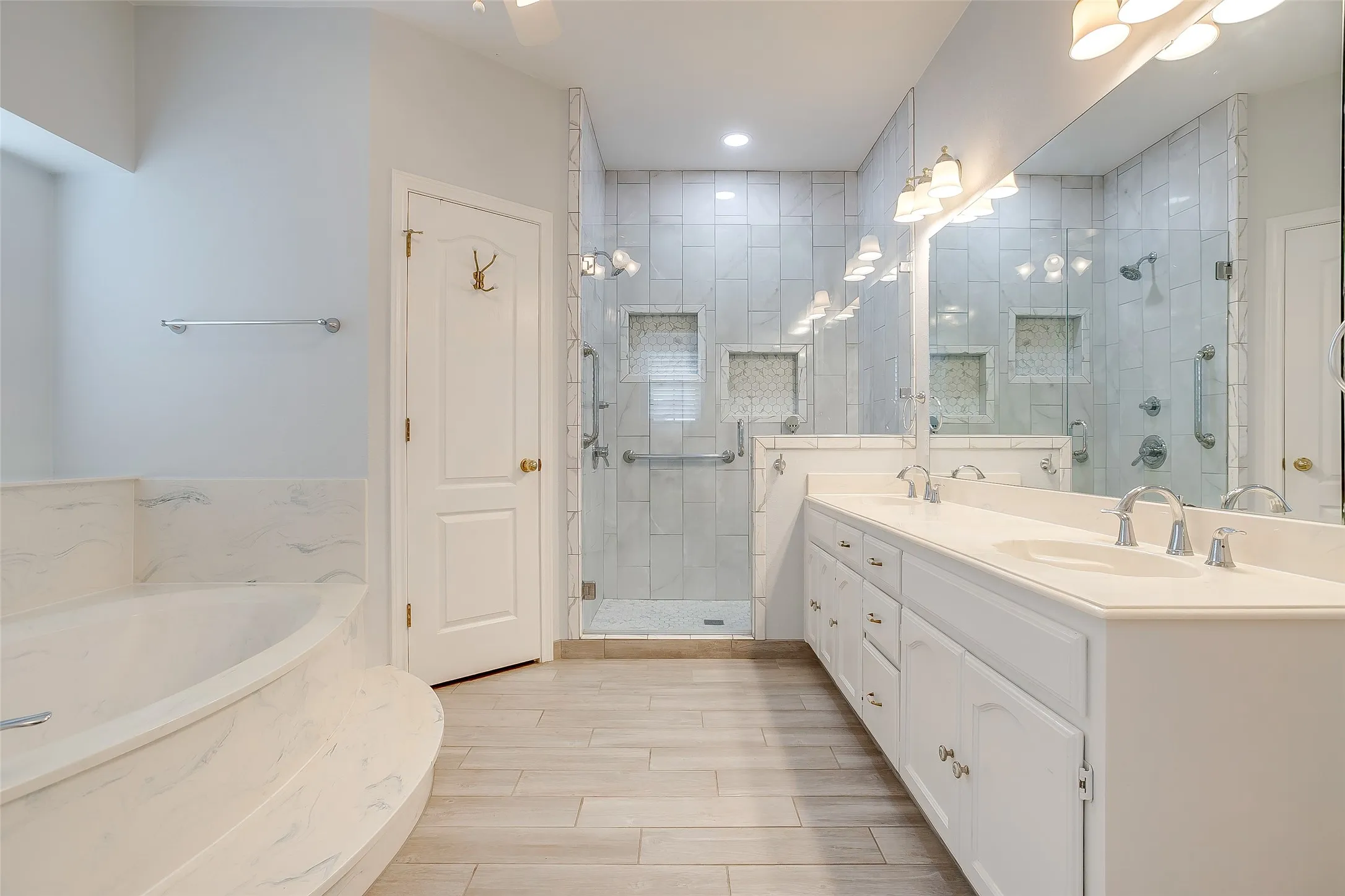 Bathroom featuring a spacious dual vanity with white cabinetry, a large walk-in shower with glass enclosure and tiled walls, a built-in soaking tub with a tiled surround, and wood-finish flooring