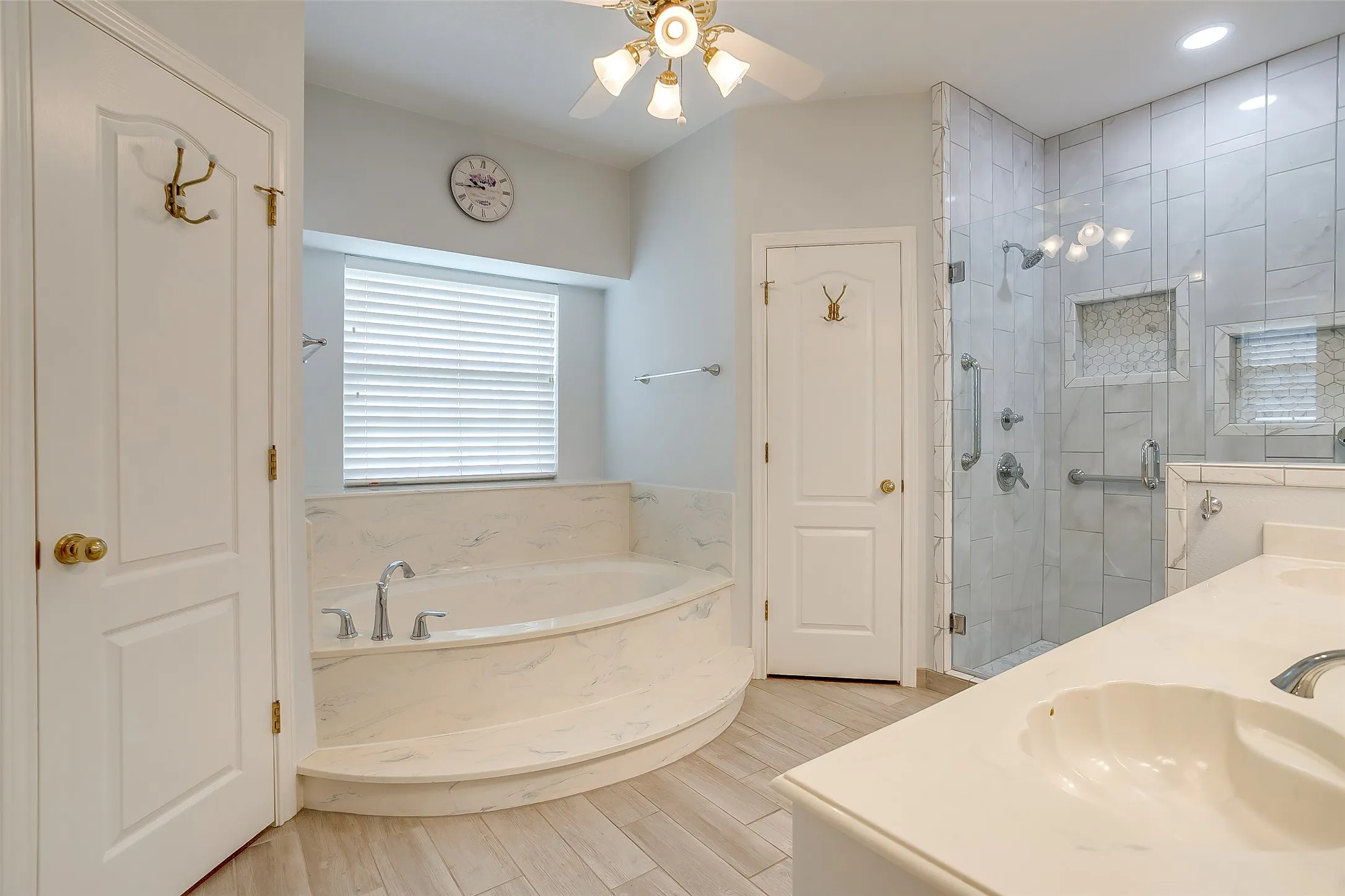 Bathroom featuring a corner soaking tub, a clear glass walk-in shower with grab bars, dual vanity with undermount sinks, wood-finish flooring, and a ceiling fan with integrated lighting