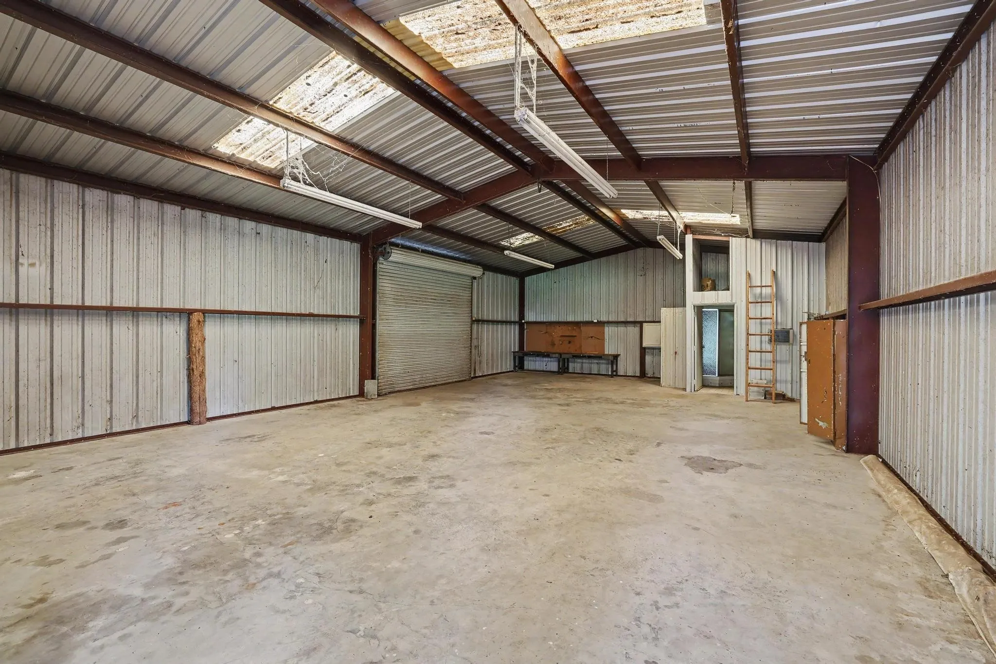 Spacious warehouse interior featuring a high metal ceiling with skylights, concrete flooring, and corrugated metal walls. Water and electricity not connected to shop.