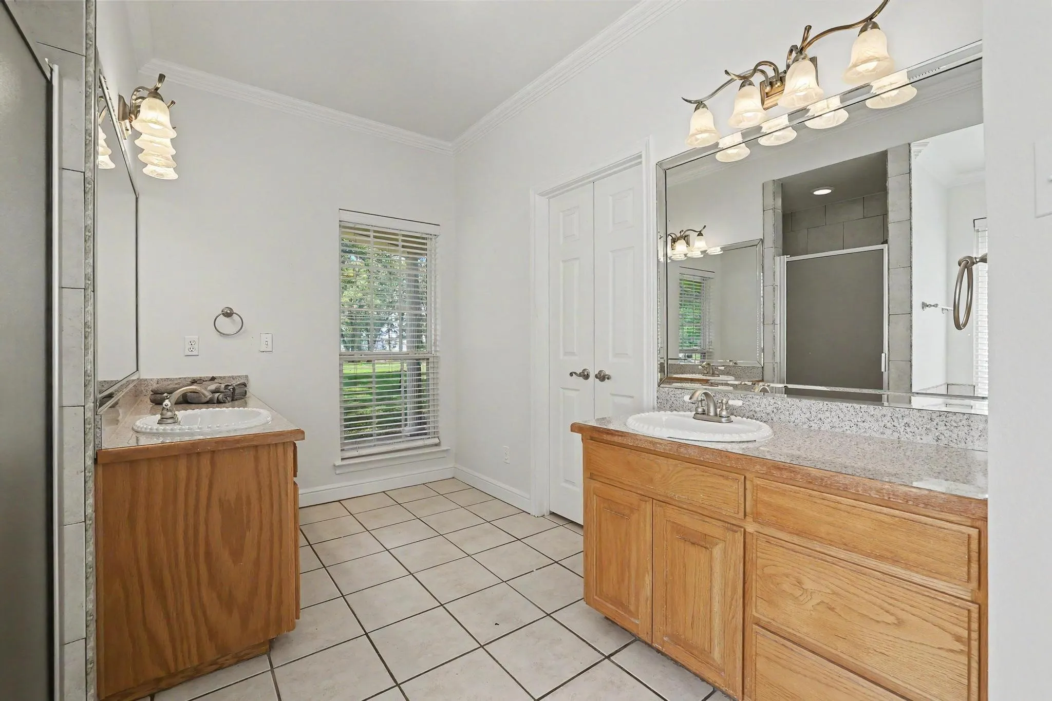 Spacious bathroom featuring dual wood-finish vanities with integrated sinks and stone-finish countertops, overhead vanity lighting, a large mirror, and tiled flooring. Through the double doors is a nicely sized walk-in closet.
