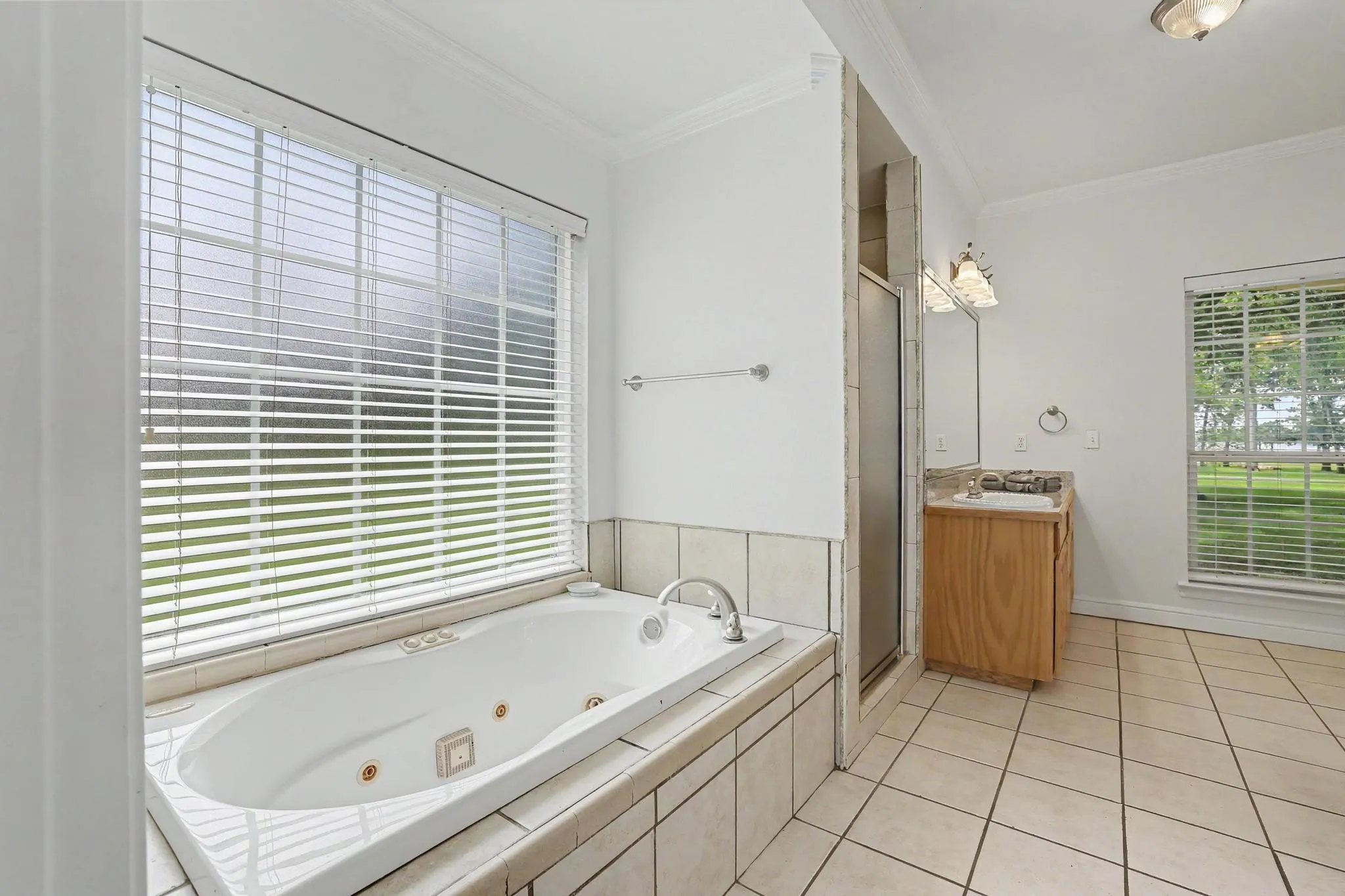 Integrated jetted tub with tiled surround, large frosted window with blinds, separate shower enclosure with frosted glass, 1 of 2 wood-finish vanities with countertop sink, and tiled flooring.