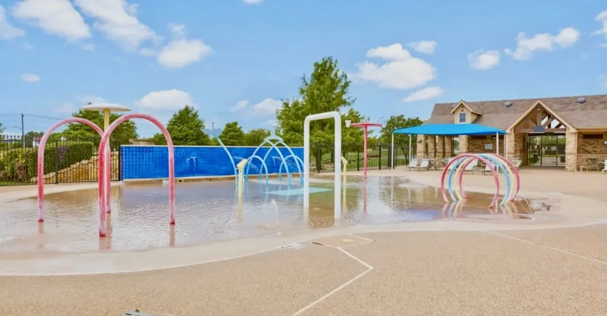 Splash pad featuring multiple water play structures, a textured ground surface, a stone-clad building with covered patio, and mature landscaping