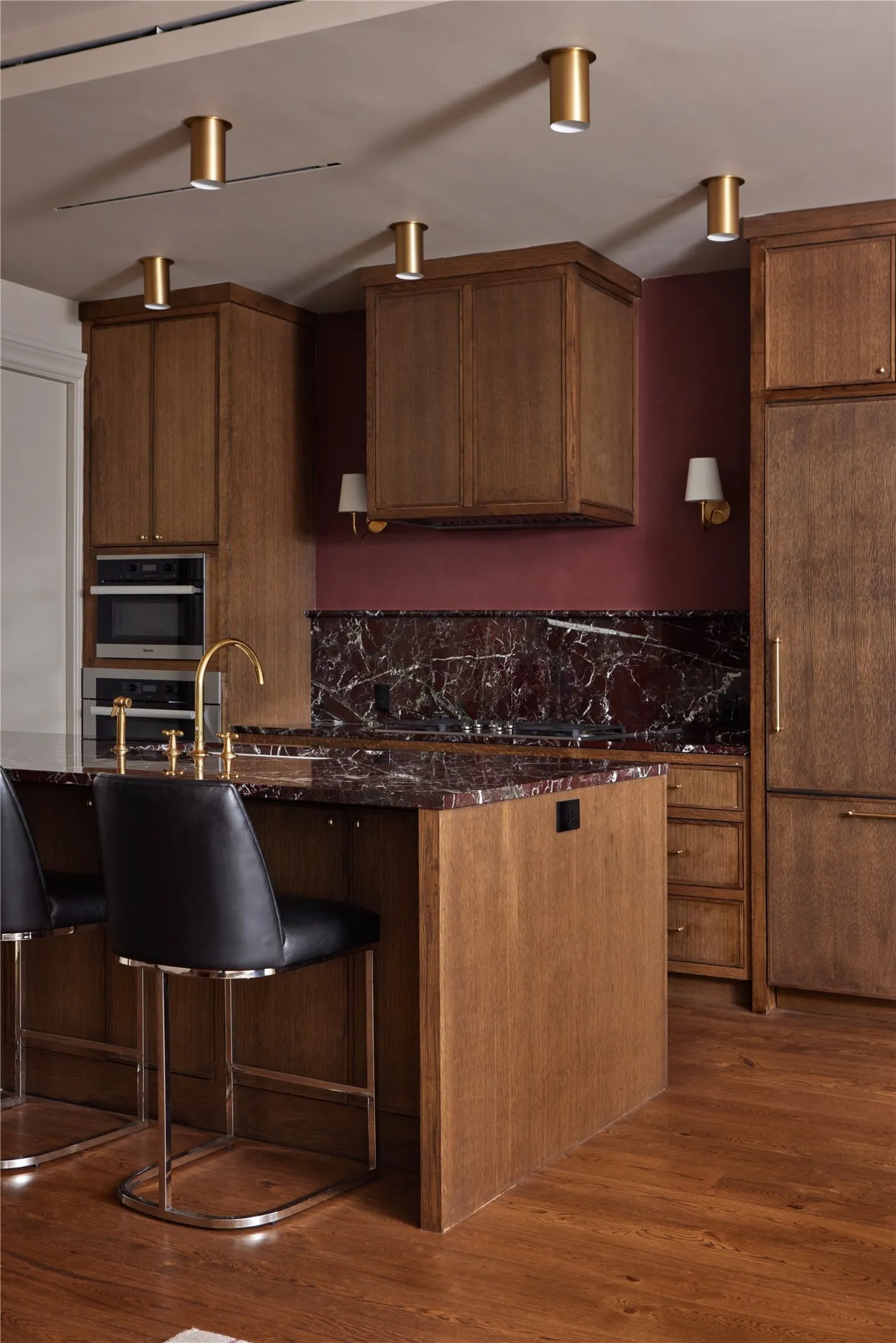 Chef's kitchen featuring rich wood cabinetry, a large center island with seating, dark veined stone countertops and backsplash, integrated stainless steel appliances, and wood-finish flooring