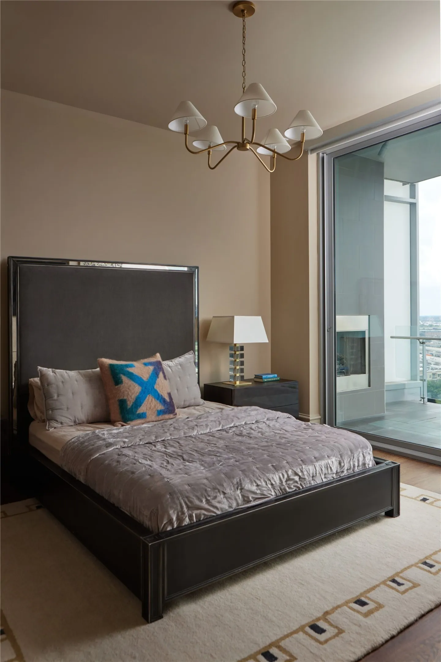 Spacious room featuring a dark-toned bed frame with a high headboard, direct access to an exterior balcony through a sliding glass door, a contemporary chandelier, and a built-in fireplace feature on the balcony