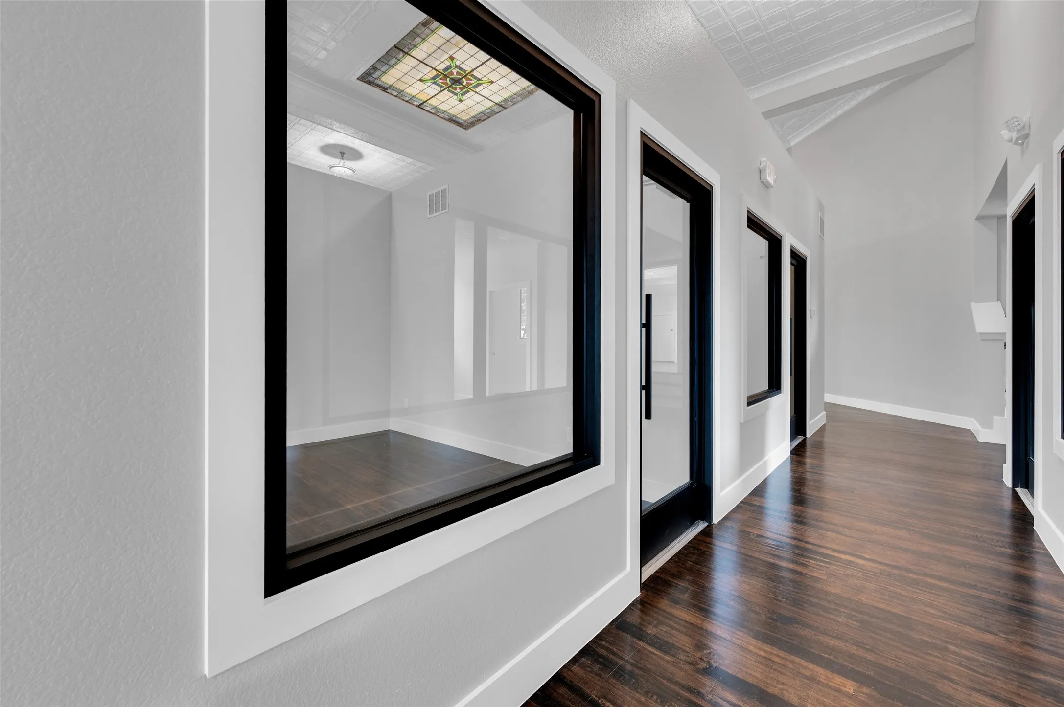 Hallway featuring rich wood-finish flooring, vaulted ceilings, and a framed internal window offering a glimpse of a room with a decorative stained-glass ceiling insert