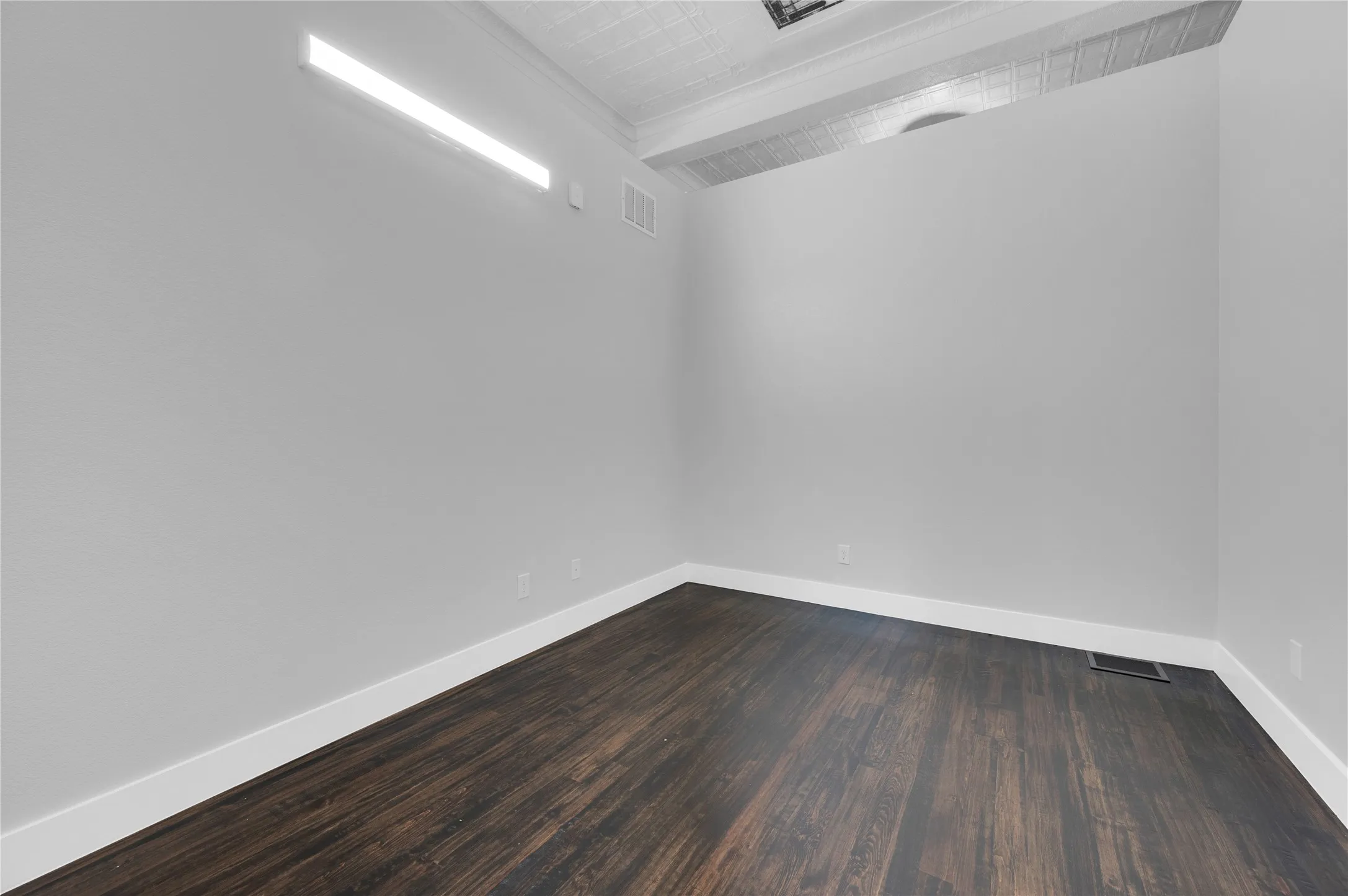 Spacious room featuring dark wood-finish flooring, white baseboards, light gray wall paint, a linear wall light fixture, and an exposed ceiling beam