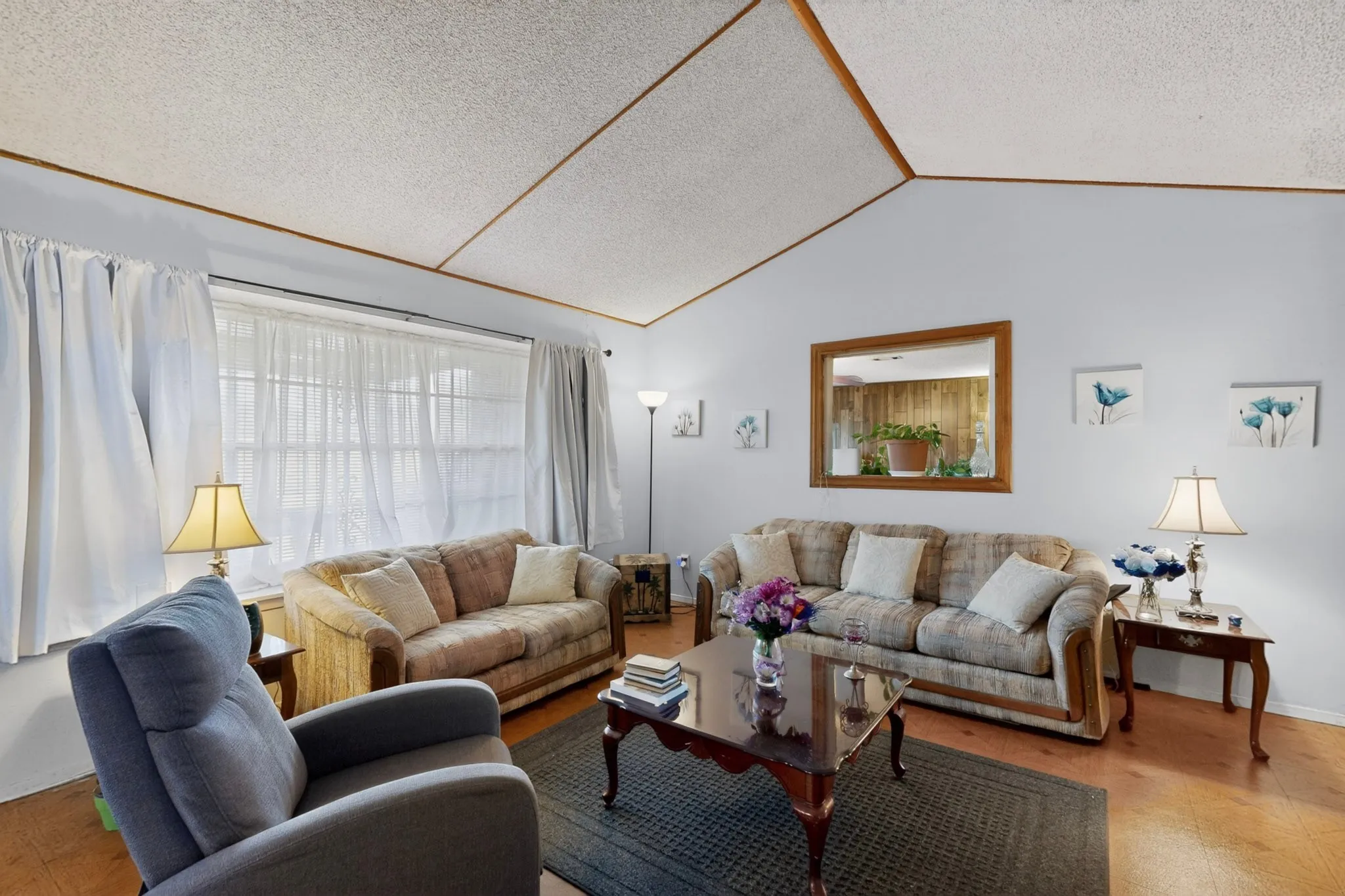 Spacious living area featuring a vaulted ceiling with exposed wood beams, hardwood flooring, and a large window with sheer and solid white curtains