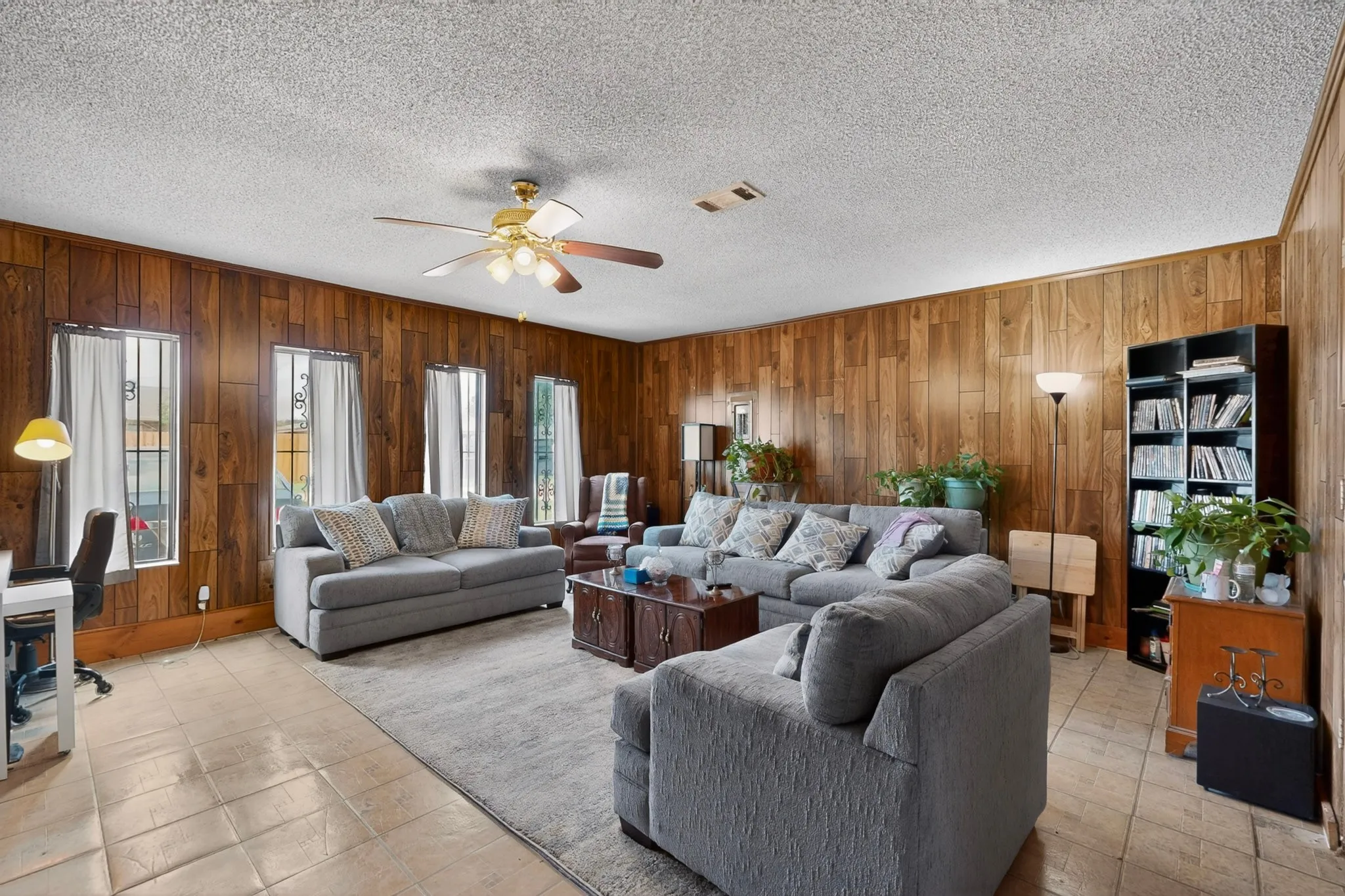Spacious interior featuring wood-panel walls, multiple windows with security bars, a ceiling fan with integrated lighting, and tile flooring