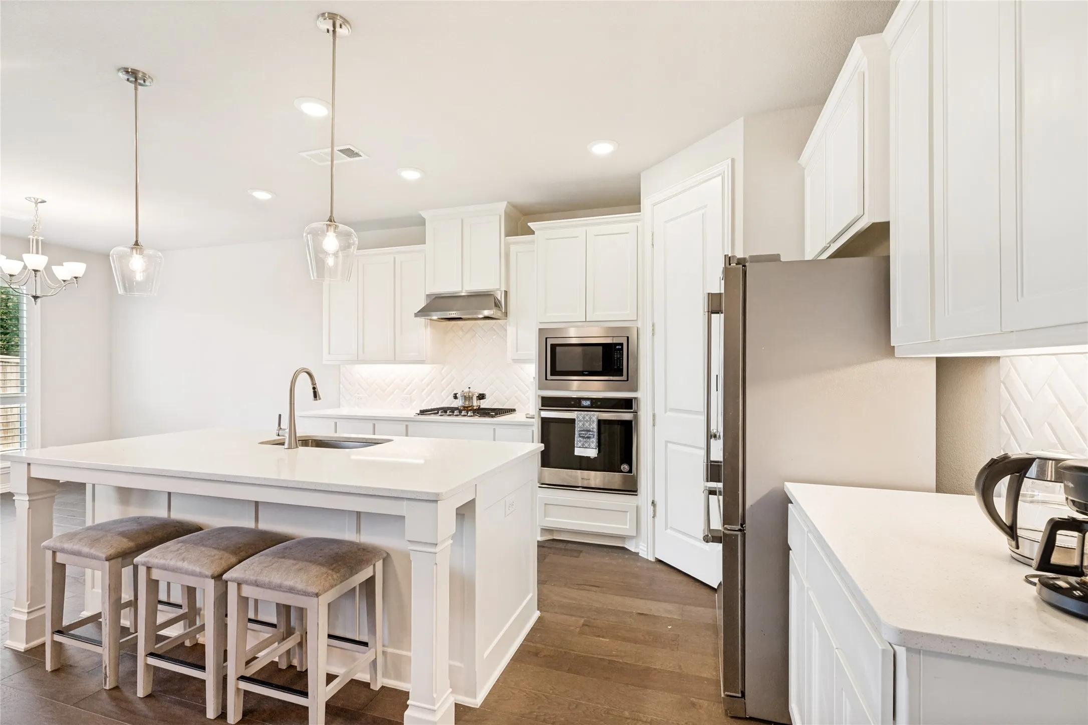 Modern kitchen featuring a large central island with integrated sink, white cabinetry, integrated stainless steel appliances, herringbone tile backsplash, and dark wood-finish flooring