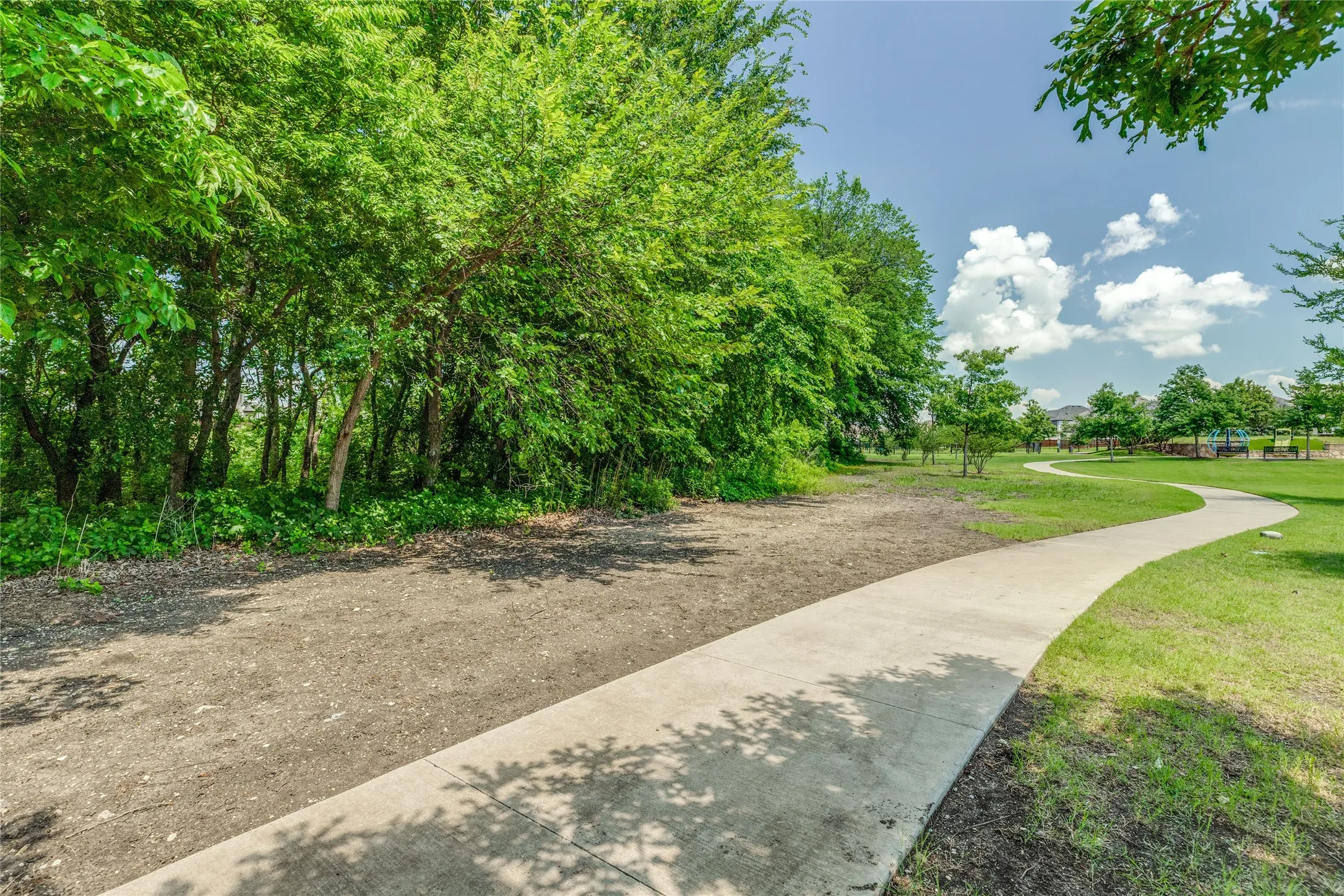 Winding concrete pathway traversing manicured green space, bordered by mature trees and natural landscaping