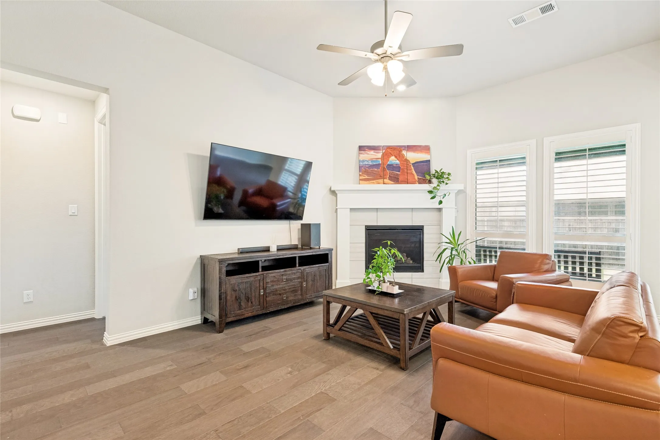 Spacious living area featuring wood-finish flooring, a fireplace with a white mantel, and two windows with horizontal blinds