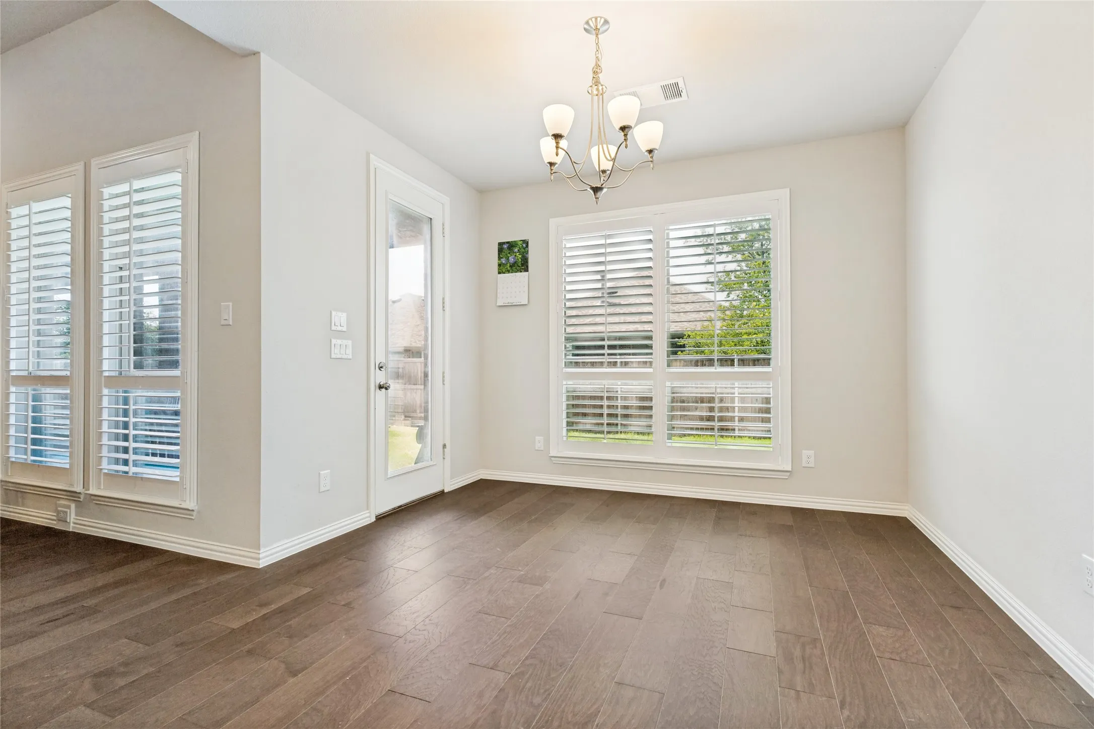 Open-concept room featuring wood-finish flooring, a traditional chandelier, multiple windows with plantation shutters, a glass-paned door, and white baseboards