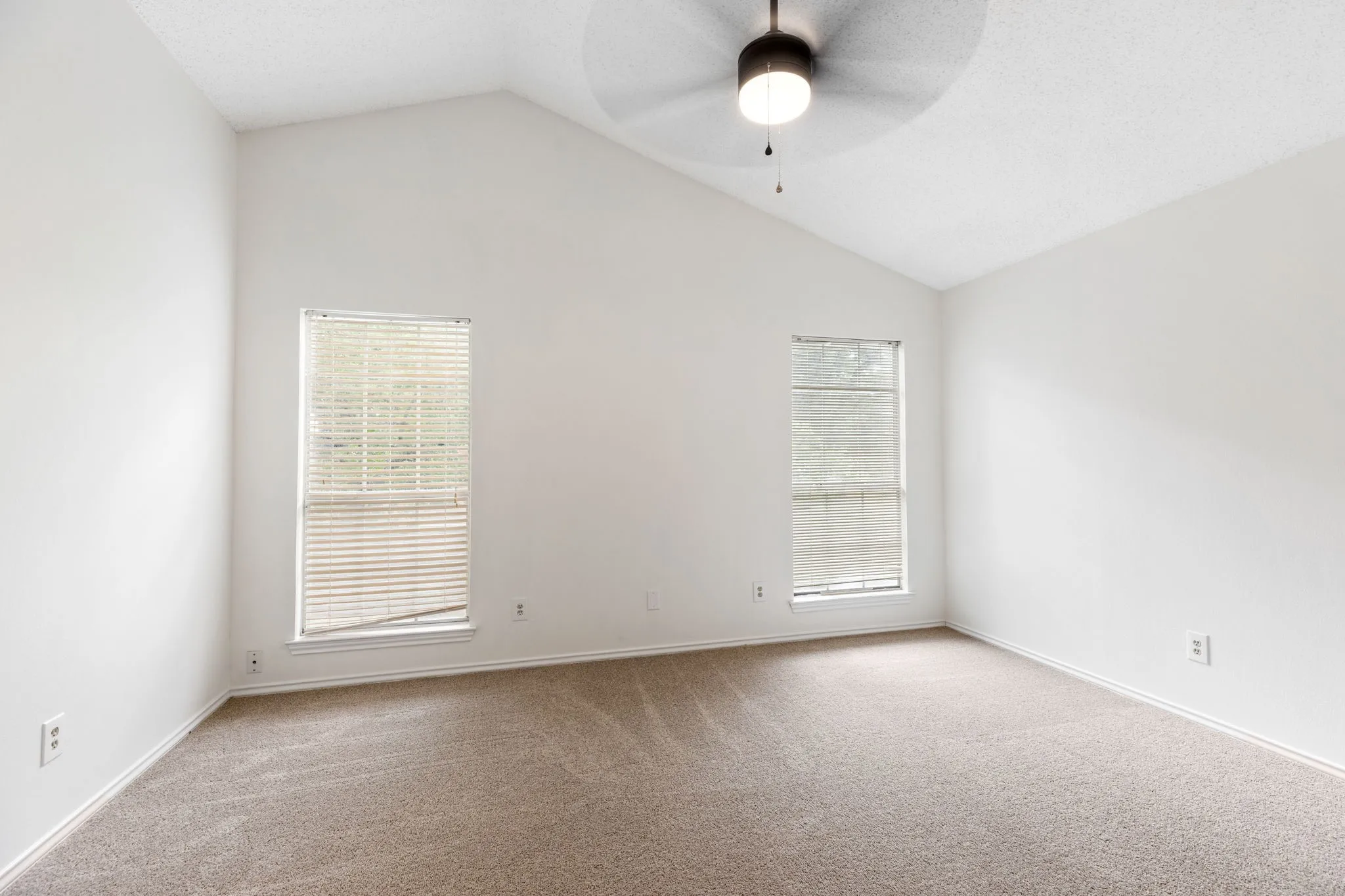 Vaulted ceiling with a ceiling fan light fixture, two windows with blinds, and neutral tone carpet flooring