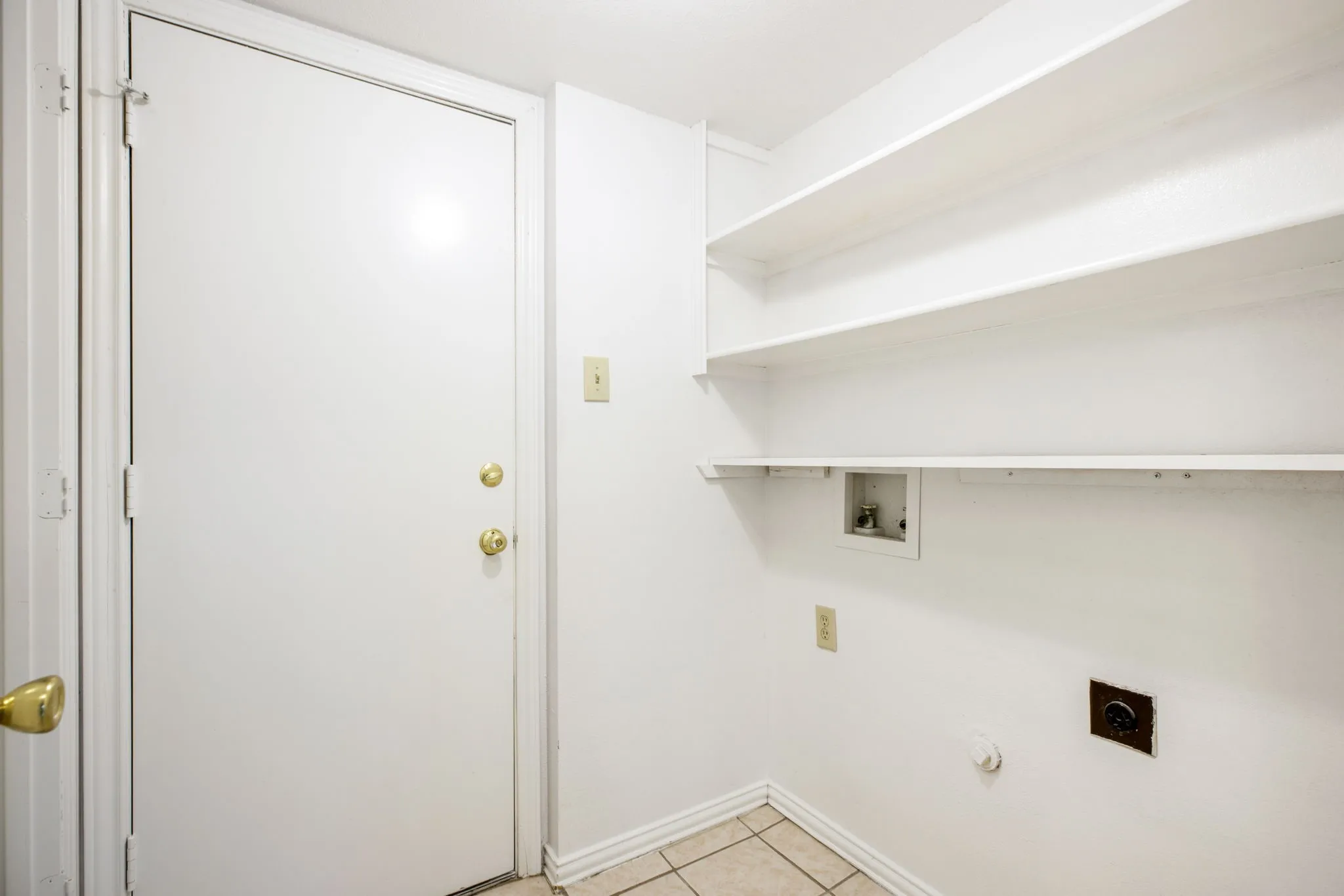Utility room featuring built-in shelving, tile flooring, a wall-mounted light switch, and a white interior door with brass hardware