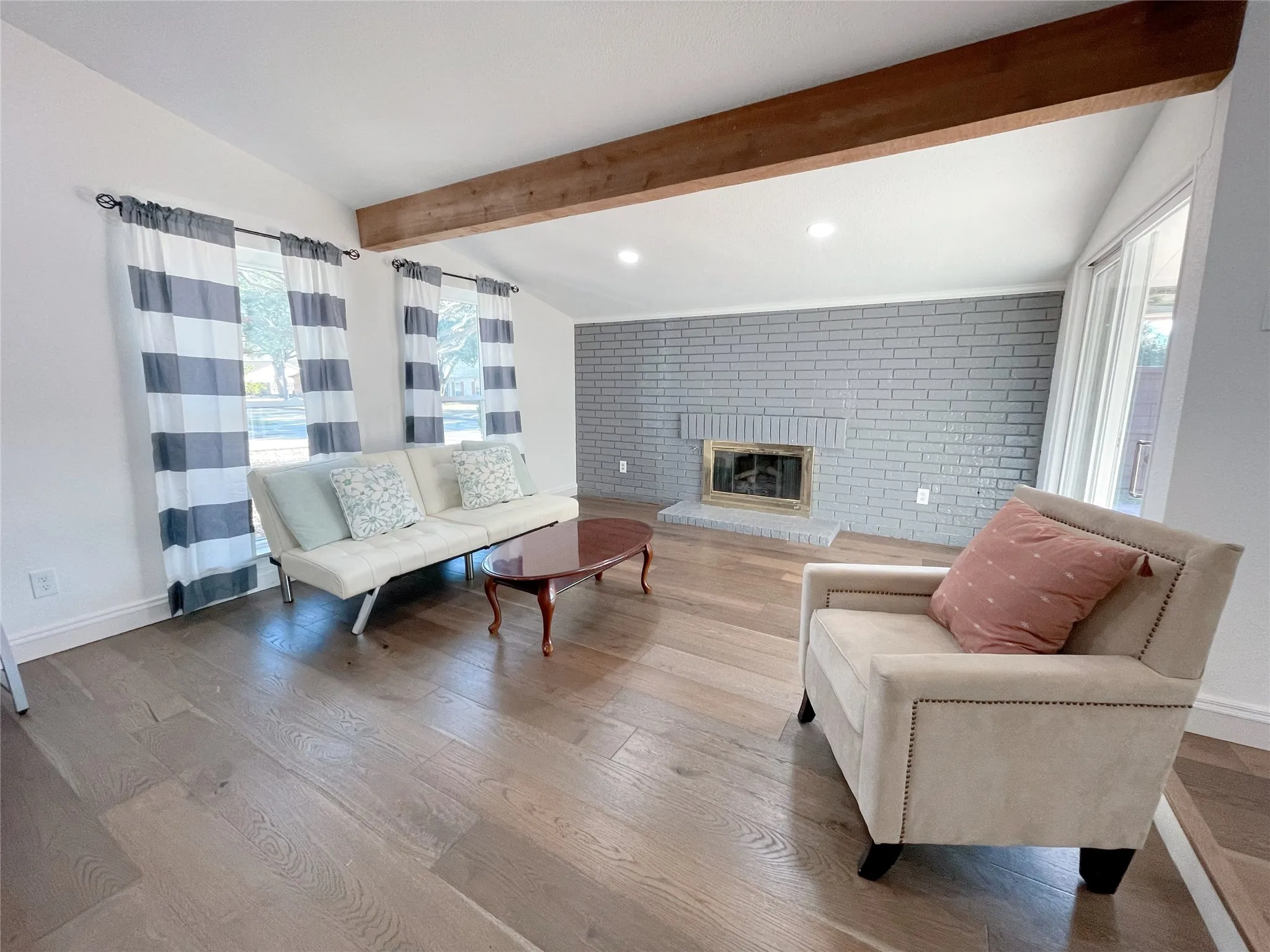Living area featuring light wood-finish flooring, a painted brick fireplace, a high vaulted ceiling with an exposed wood beam, recessed lighting, and white baseboards