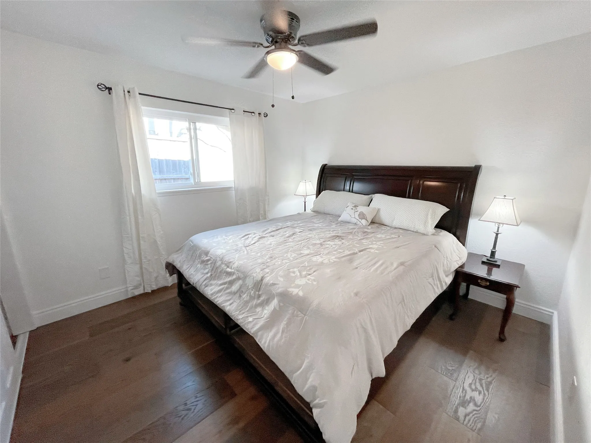 Spacious room featuring wood-finish flooring, a ceiling fan with integrated lighting, a large window, and crisp white walls
