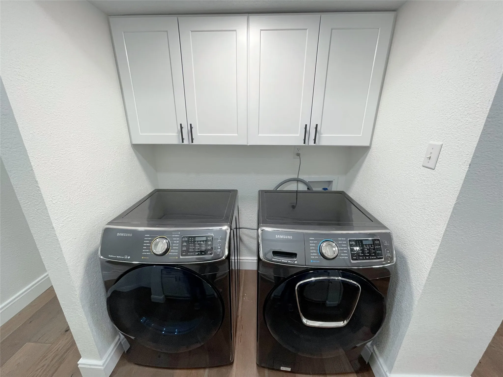 Dedicated laundry area featuring overhead white shaker cabinets with matte black hardware, wood-finish flooring, and white baseboards