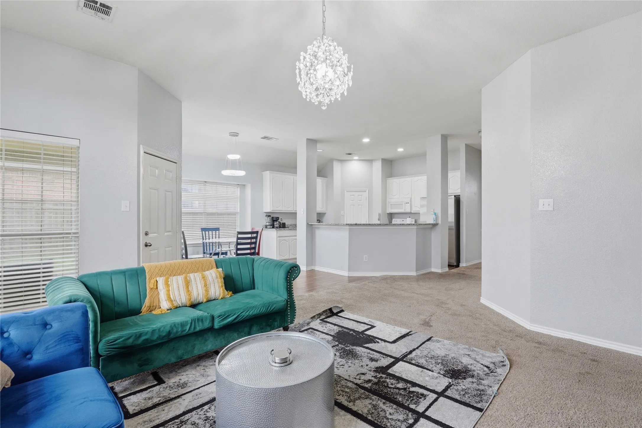 Spacious open-concept living area featuring light-toned walls, neutral carpeting, and a prominent crystal chandelier