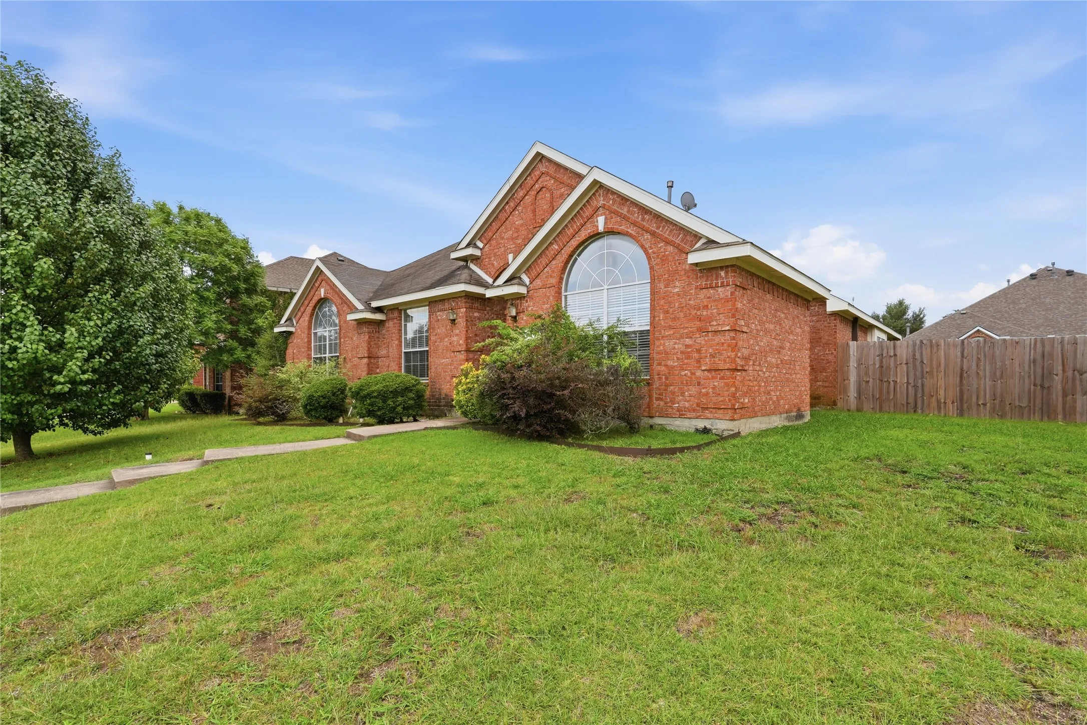 Brick exterior with an arched window, mature trees, a manicured lawn, and a wooden privacy fence