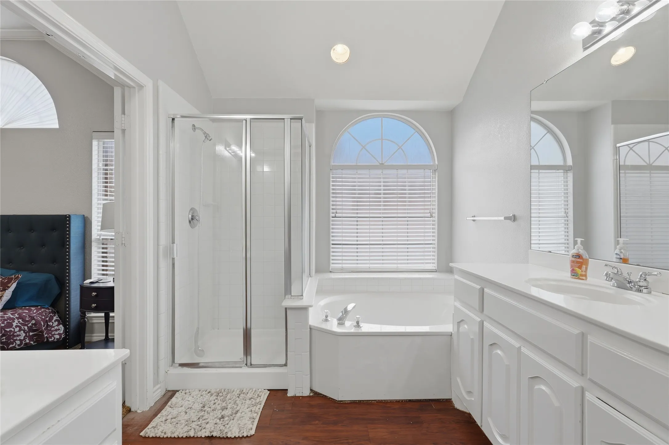 Bathroom featuring a built-in corner soaking tub with arched window, a separate glass-enclosed shower, wood-finish flooring, and a double vanity with a large mirror