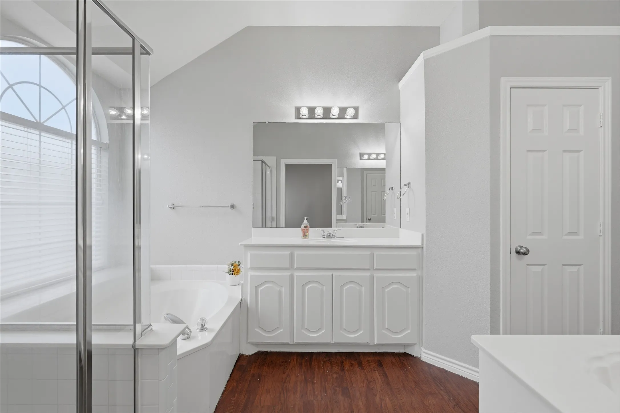 Bathroom featuring a corner soaking tub, glass-enclosed shower, wood-finish flooring, a white vanity with an integrated sink, and a large mirror with overhead lighting