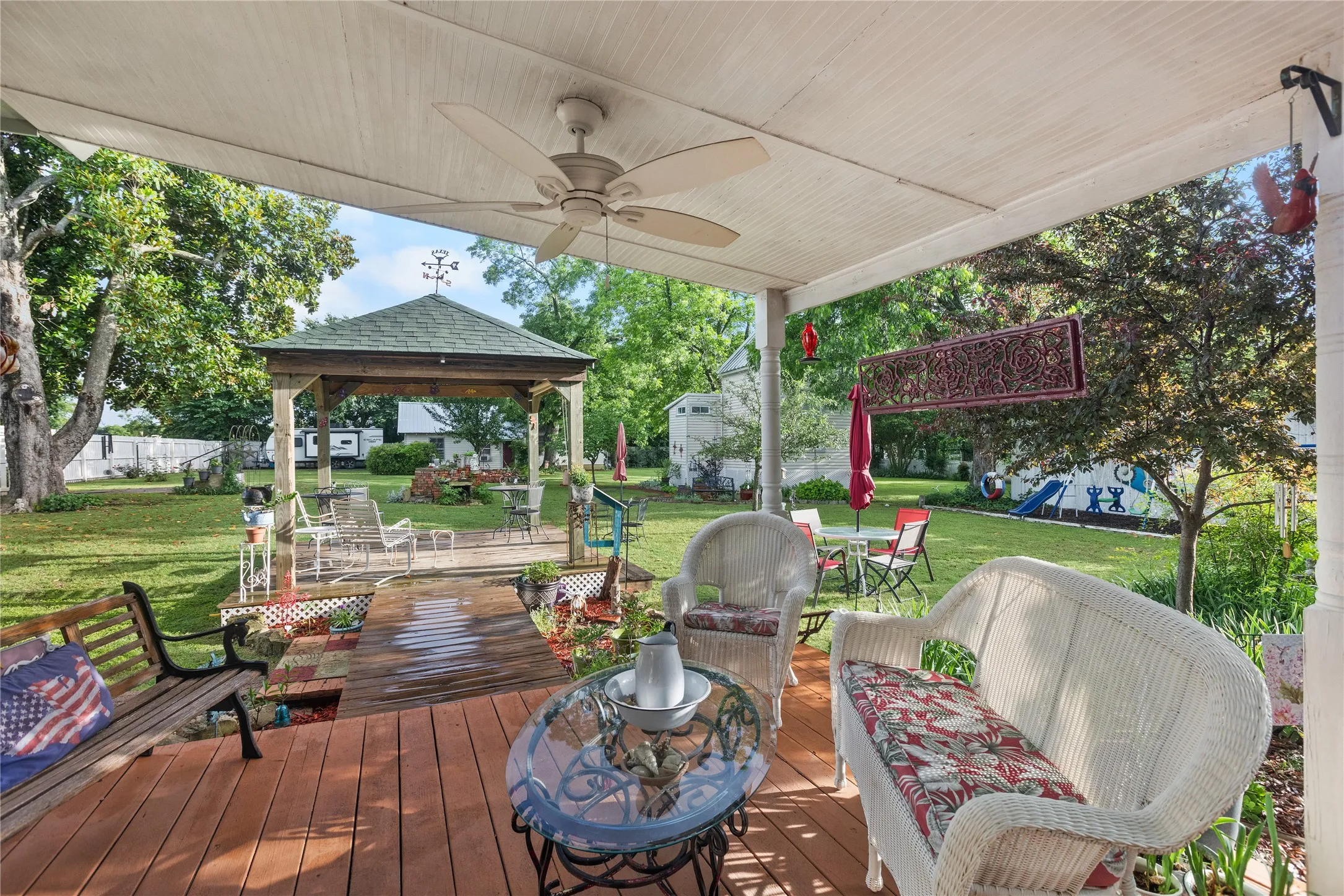 Covered porch with wood-finish flooring, featuring a ceiling fan, white support columns, and an expansive grassy yard beyond