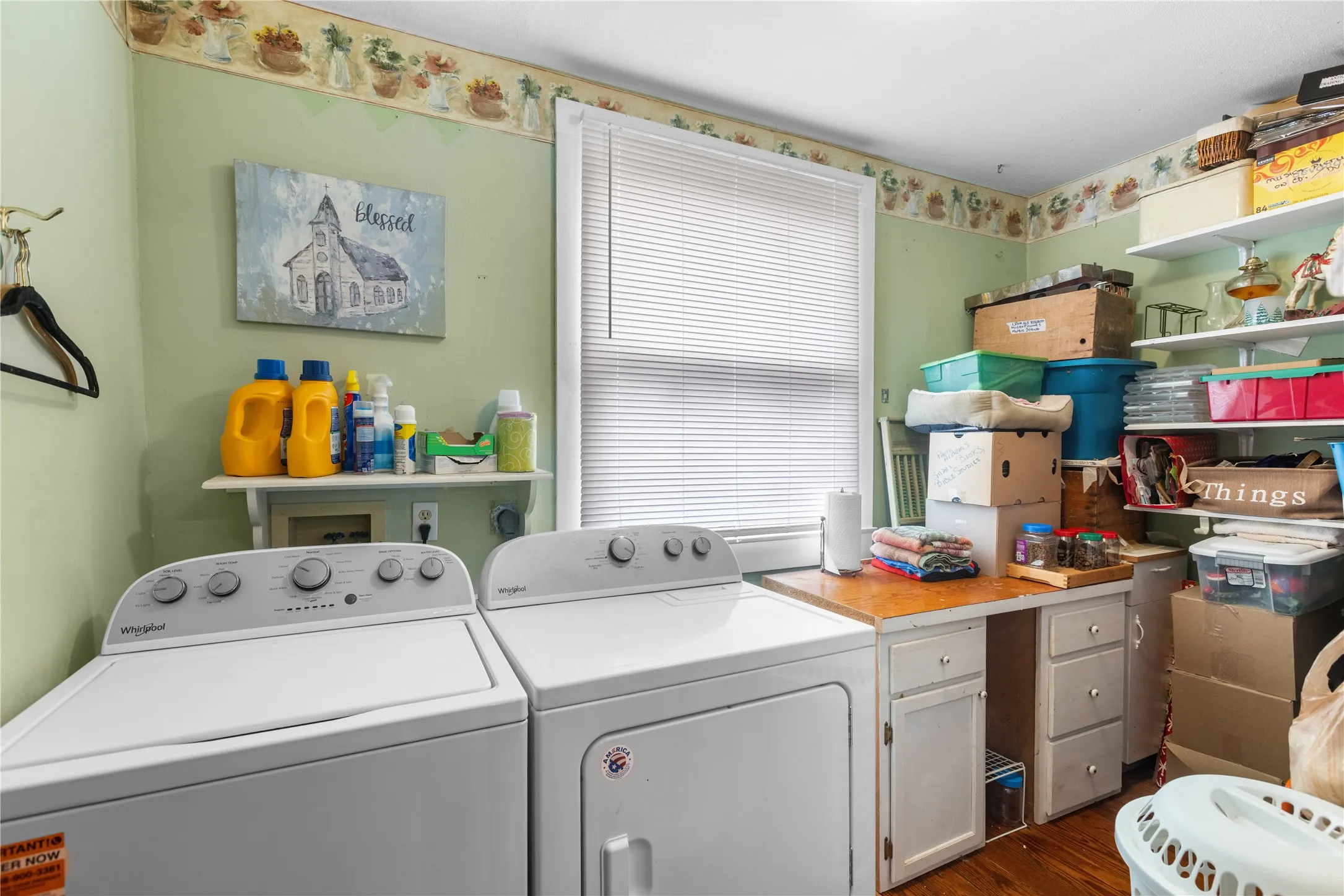 Dedicated laundry area featuring wood-finish flooring, a bright window with blinds, a built-in countertop, and wall-mounted shelving